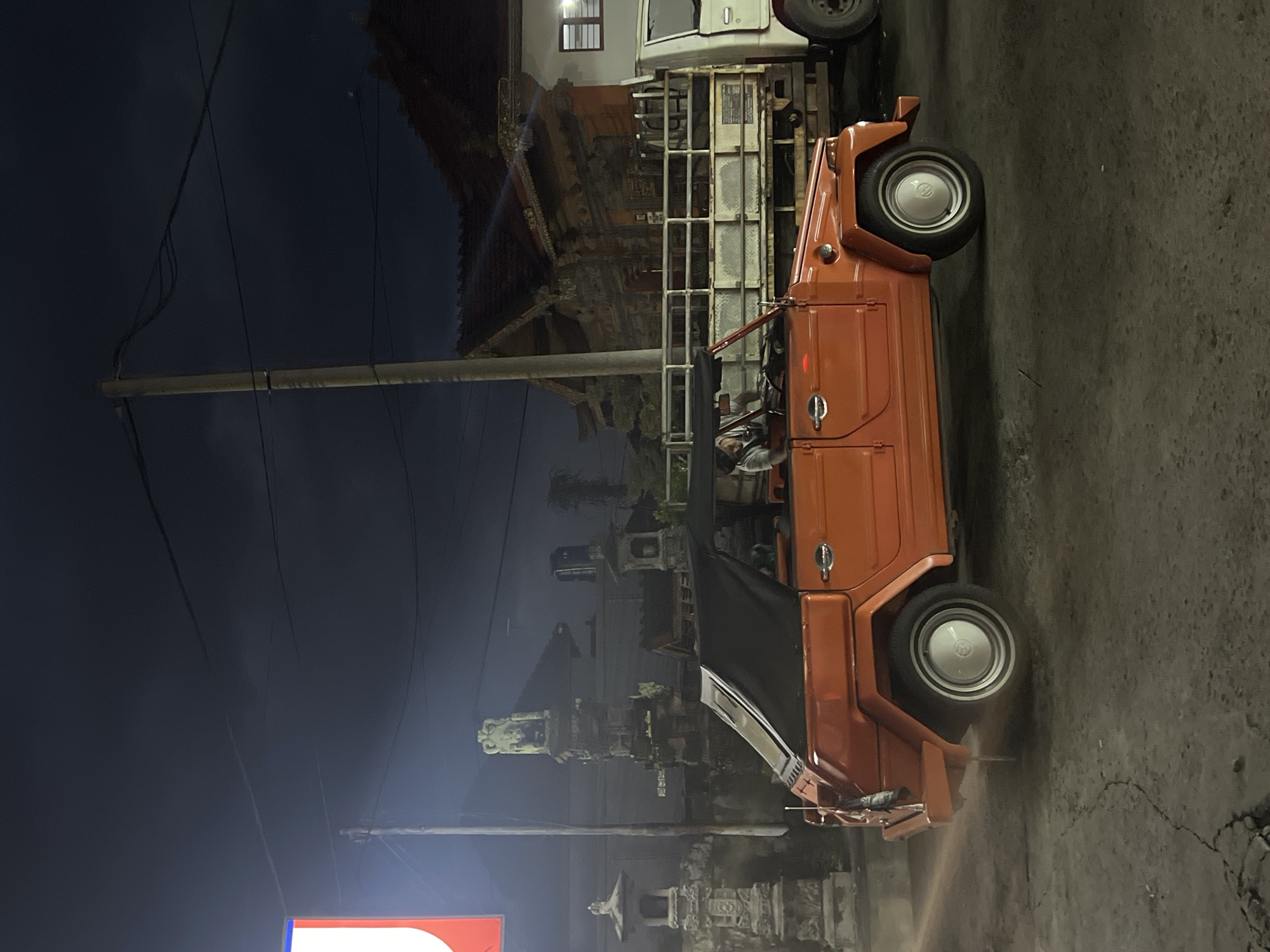 A photograph captures a roadside scene in Sukawati, Indonesia, at night. The dominant subject is an orange Volkswagen Thing vehicle parked on the right side of the frame, facing upward. A white van or truck is partially visible above the Volkswagen. To the left of the vehicles, there are structures resembling Balinese shrines or small temples. A tall utility pole stands at the left edge of the frame, with power lines stretching across the dark sky. The pavement shows signs of wear and cracking. A sign with a red and white design appears at the bottom-left corner of the frame. The overall lighting suggests it is nighttime, with artificial lights illuminating the street and structures. There are at least two occupants inside the orange vehicle.