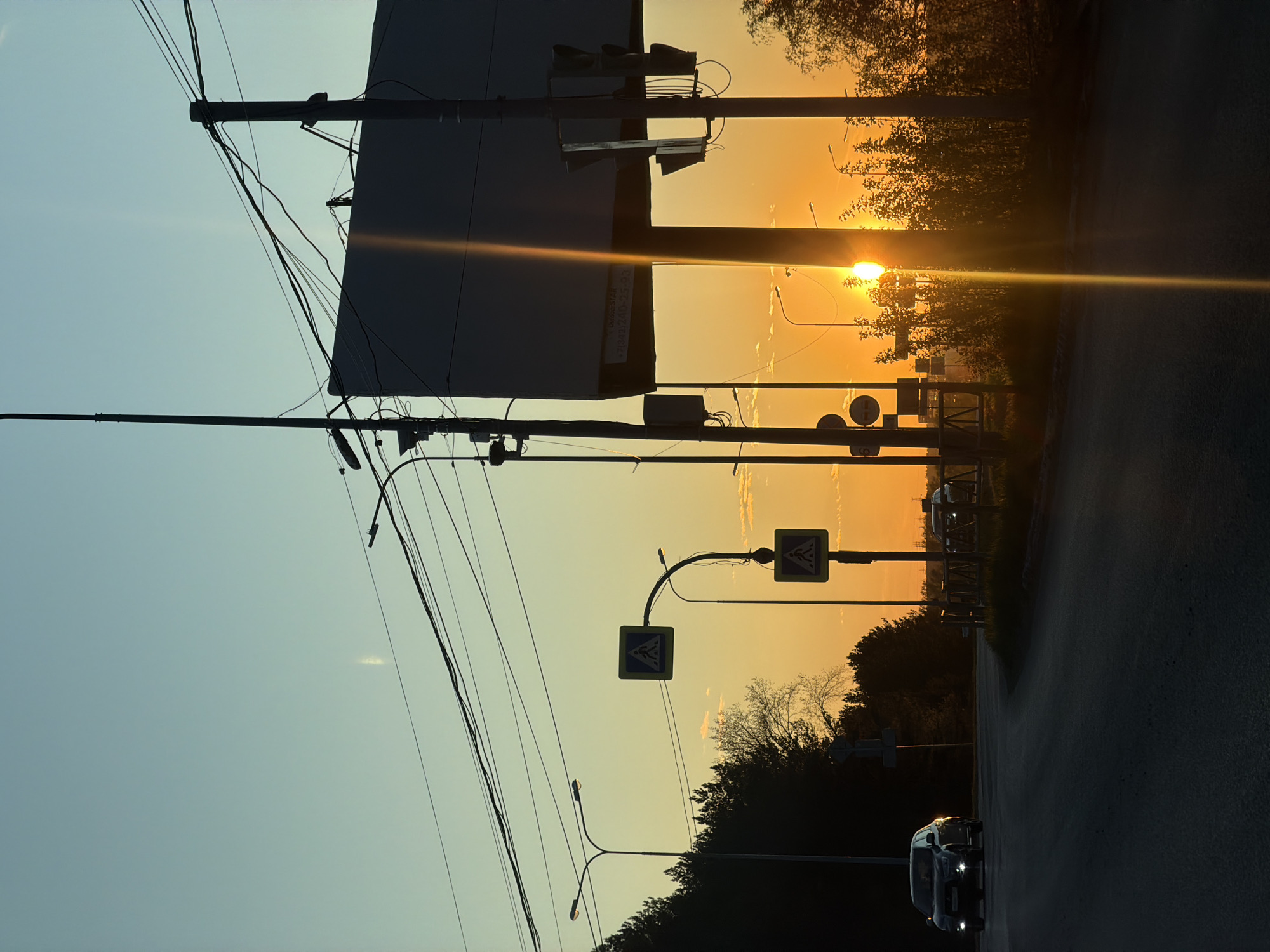 The image shows a street in Chaikovsky, Russia at sunset. The sky transitions from blue to orange as the sun dips below the horizon. Silhouetted against the sky are power lines, a large billboard, traffic lights, pedestrian crossing signs, street lamps, and trees. A car with headlights on is traveling on the road towards the camera. The asphalt road is dark.