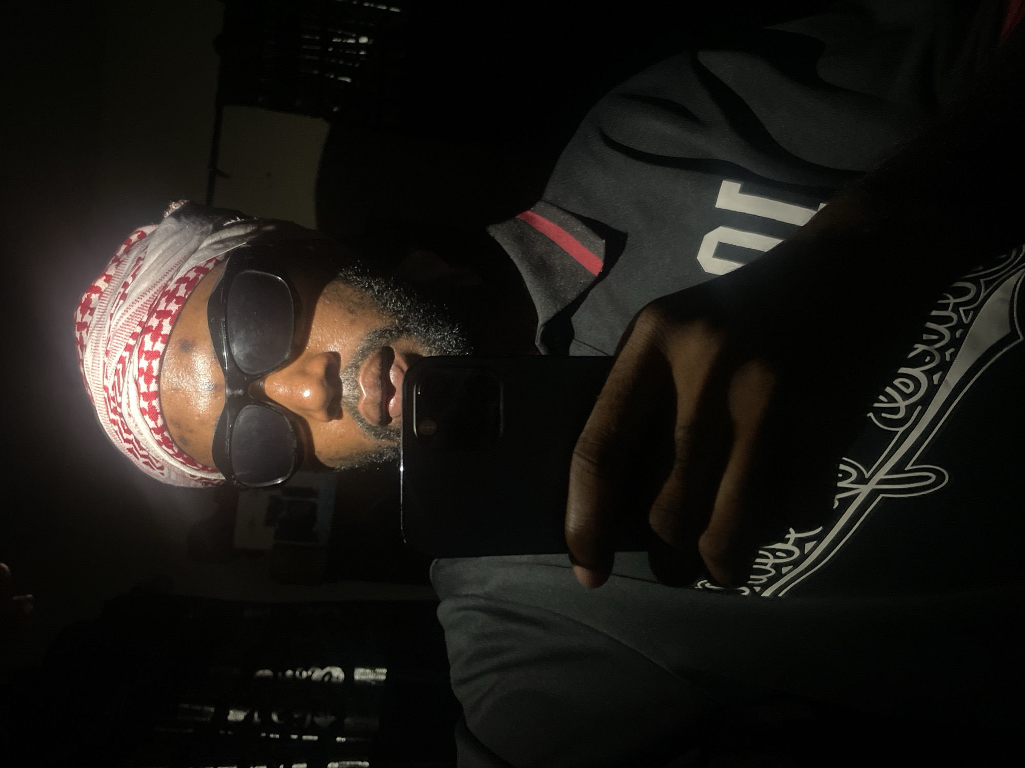 The image is of a man, likely taking a selfie, in a dark environment. The man is wearing a red and white patterned head covering, sunglasses, and a dark-colored garment with a white paisley-like design. He is holding a black smartphone. Given the darkness and lack of environmental context, it is impossible to determine a specific location in Lokoja, Nigeria, or even confirm if the image was taken there.