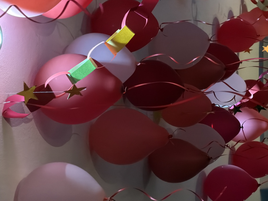 A close-up, upward-angled view captures numerous helium-filled balloons and paper decorations suspended against a light-colored ceiling. The scene is located in San Bernardino Tlaxcalancingo, Mexico. Balloons are predominantly in shades of red, ranging from light pink to dark maroon, with several white balloons interspersed. Each balloon is affixed with a curled red ribbon, extending downwards. Overhead decorations include sections of a paper chain, featuring green and yellow links, from which golden star-shaped cutouts are suspended by thin strings. Shadows cast by the balloons and decorations are visible on the ceiling, indicating an indoor environment with overhead lighting. No individuals or specific actions are depicted.
