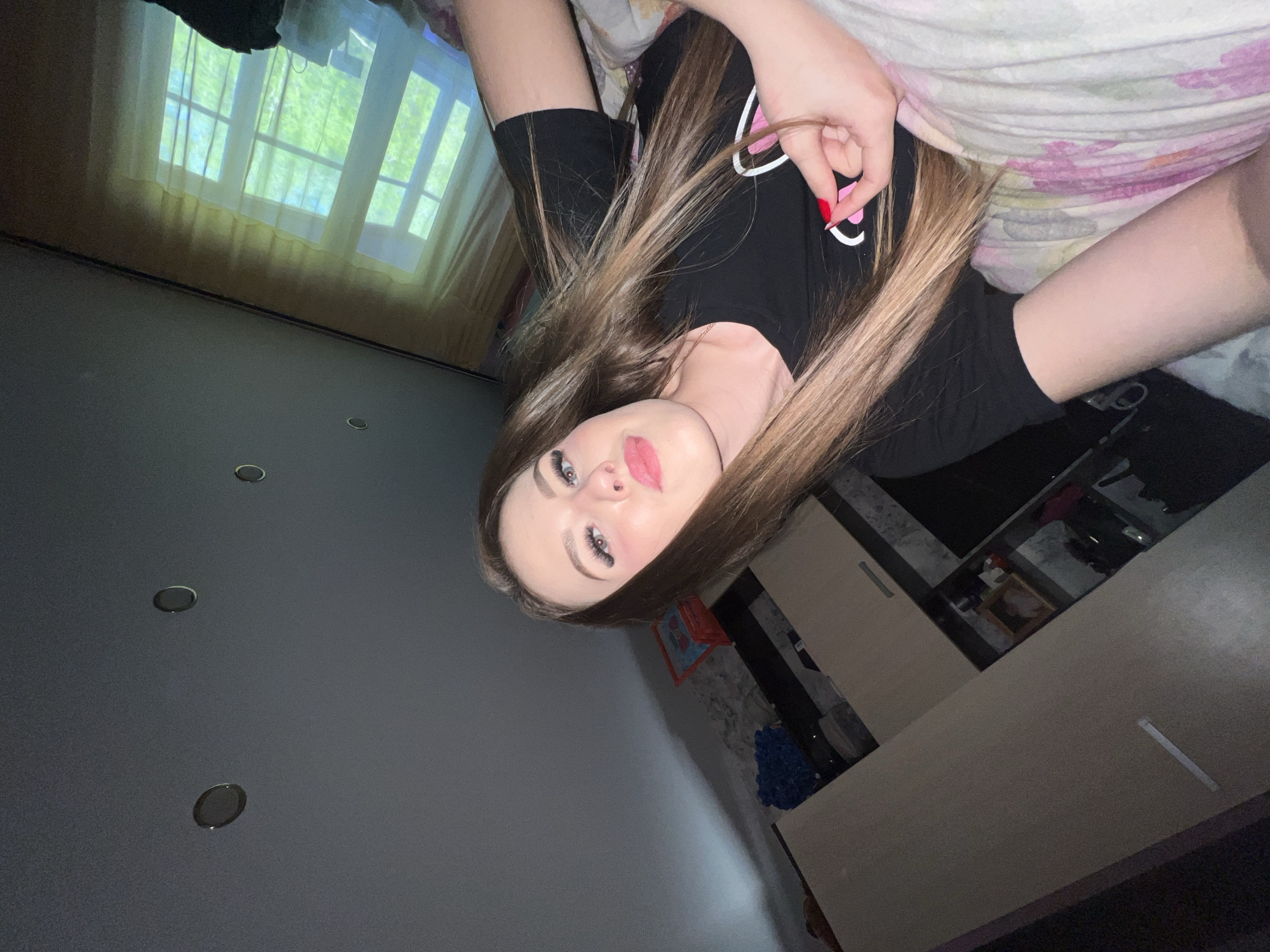 A young woman with long brown hair and pink lipstick is lying down, possibly on a bed, wearing a black t-shirt. Her right hand, with red nail polish, is near her face. The background includes a window with sheer curtains, a gray wall with three recessed lights, and a glimpse of furniture including what appears to be light-colored cabinets or a dresser. There are no elements identifiable to the city of Noviy, Russia.