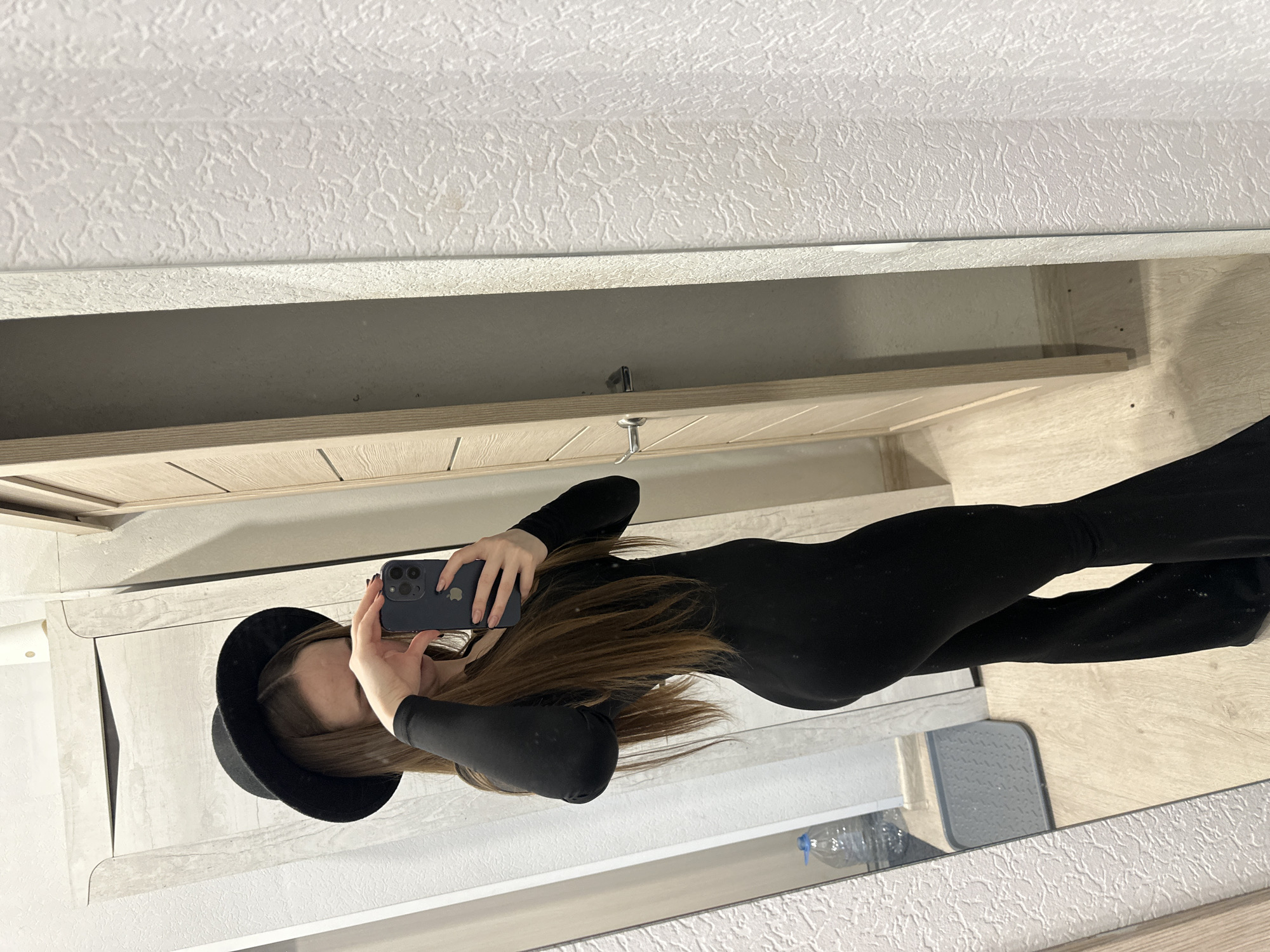 A mirror selfie shows a woman with long brown hair, wearing a black bodysuit and a black hat. She is holding a phone in both hands, presumably to take the picture. The background includes a textured wall and a wooden cabinet. The location is Unknown, Unknown.