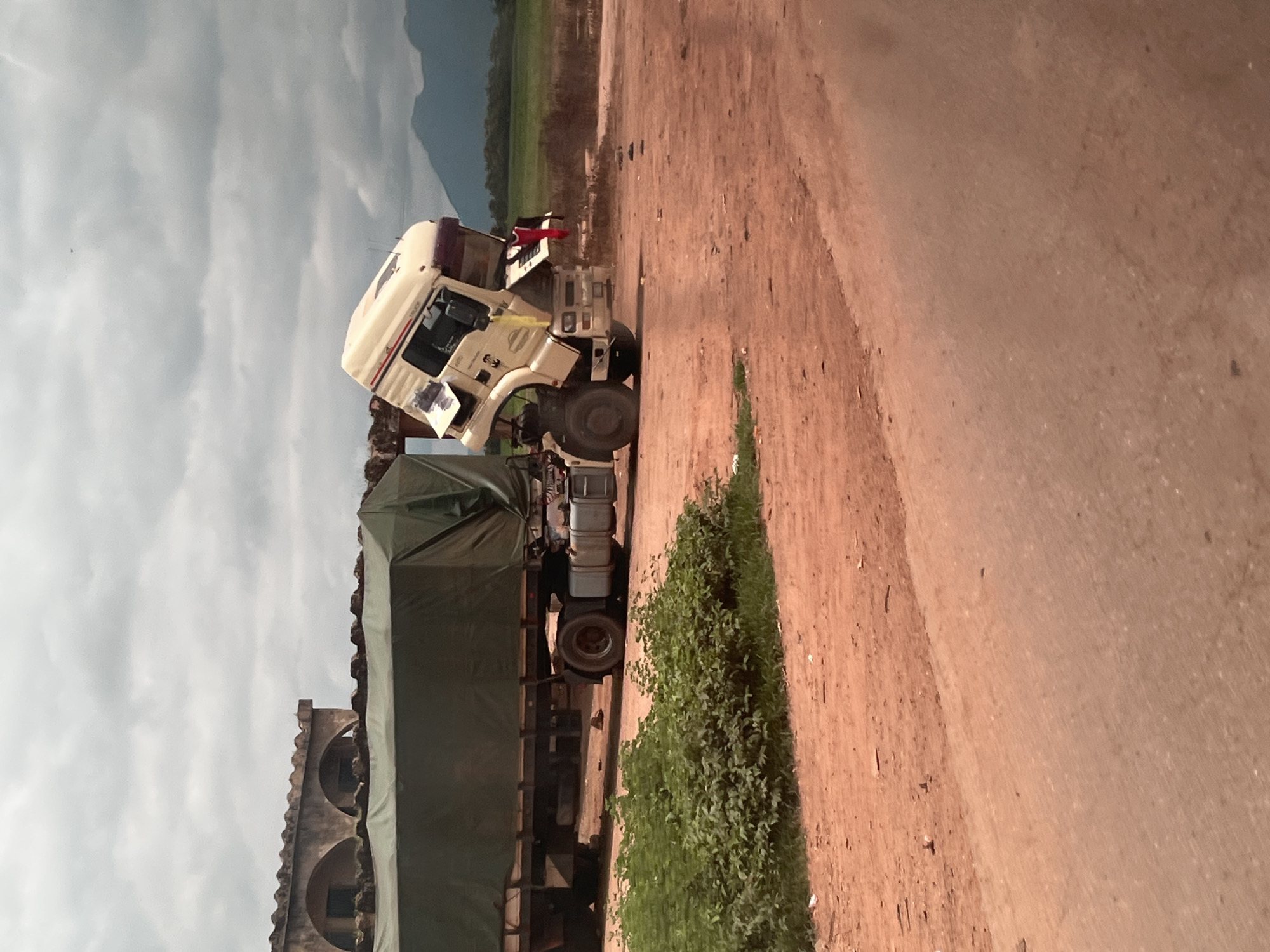 The image depicts a semi-trailer truck with its cab tilted at a severe angle, suggesting a mechanical failure or an accident. The truck is positioned on a dirt road or unpaved area, with a large, green tarp-covered trailer attached. In the background, there is a low building with arched openings and a tiled roof. Further in the distance, rolling hills are visible under a cloudy sky. The overall scene appears to be in a rural or semi-rural environment. The specific location in Karshi, Nigeria, is not visually discernible from the image itself, but the description assumes this context.