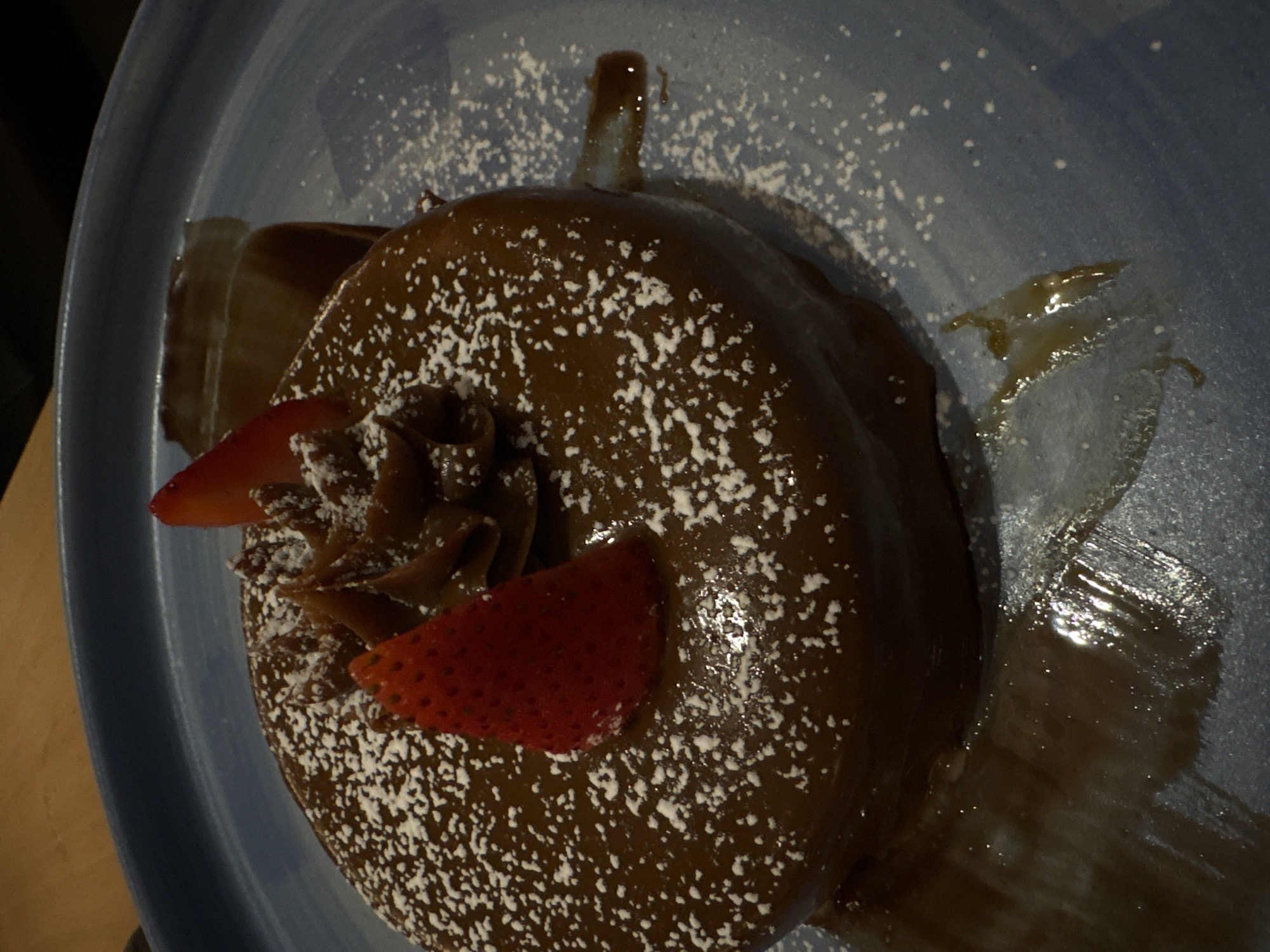 A close-up shot displays a chocolate dessert on a light blue plate, likely taken indoors. The dessert is a round, cake-like item covered in a glossy chocolate glaze and dusted with powdered sugar. Garnishing the top are two slices of fresh strawberry and chocolate ganache piped in a decorative rosette. The plate has streaks of what seems to be caramel or chocolate sauce. The photo appears to have been taken at an angle, providing a top-down view with a slight tilt. There are no people visible, and the location, while suspected to be in Mexico City, is not definitively confirmed by the image alone.