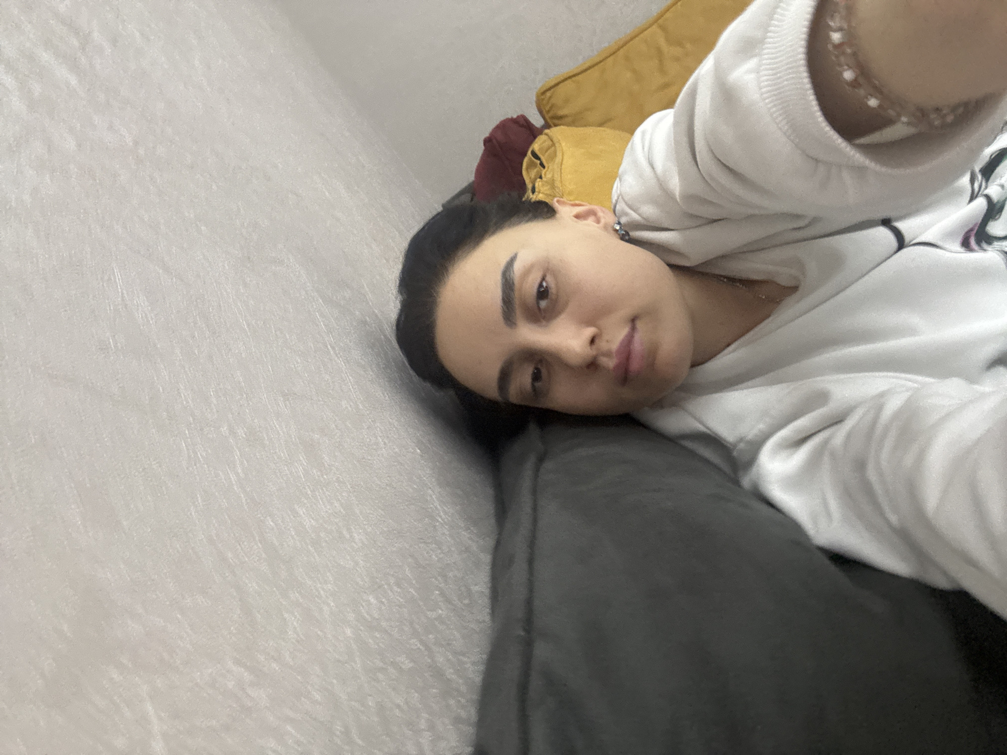 A woman is lying on a gray couch, with her head near a light gray wall. Behind her head are yellow and maroon pillows. She is wearing a white sweatshirt and a bracelet. The location cannot be determined from the image.