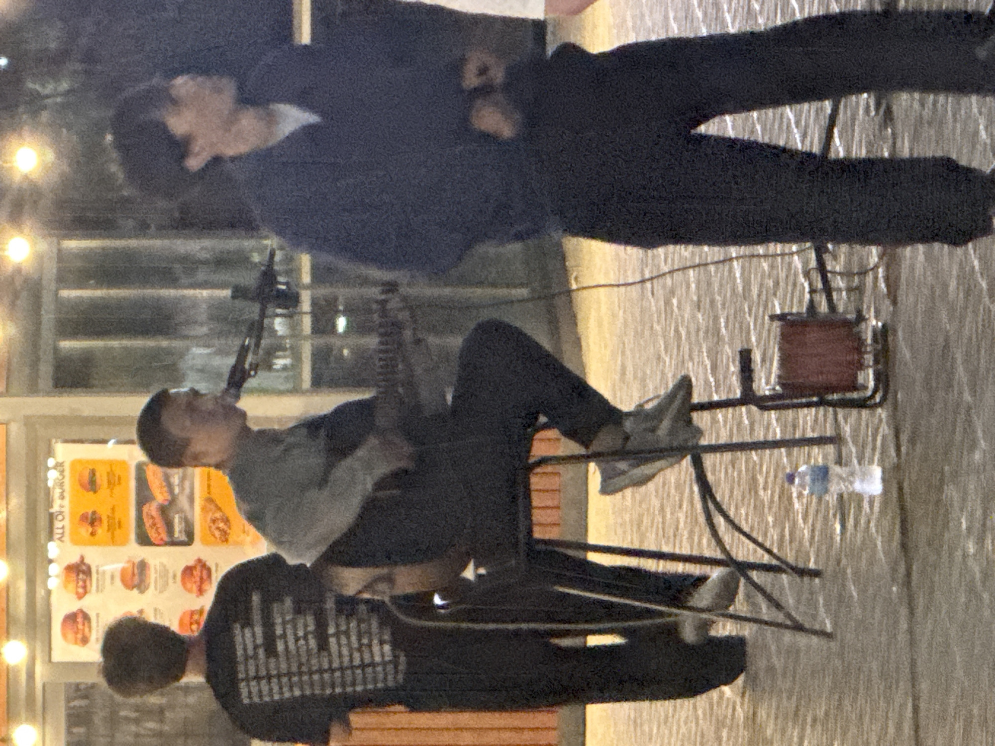 A man sits on a stool playing a guitar and singing into a microphone in front of a fast-food restaurant. Another man stands to his left wearing a black t-shirt with white text, and a third man stands to his right wearing a dark blue jacket. The restaurant has a menu displayed in the window featuring pictures of burgers and hot dogs. The ground is paved with light-colored tiles and the scene appears to be at night. The presence of the restaurant and the attire of the individuals suggest an urban or commercial setting, potentially in Pavlodar, Kazakhstan, given the prompt.