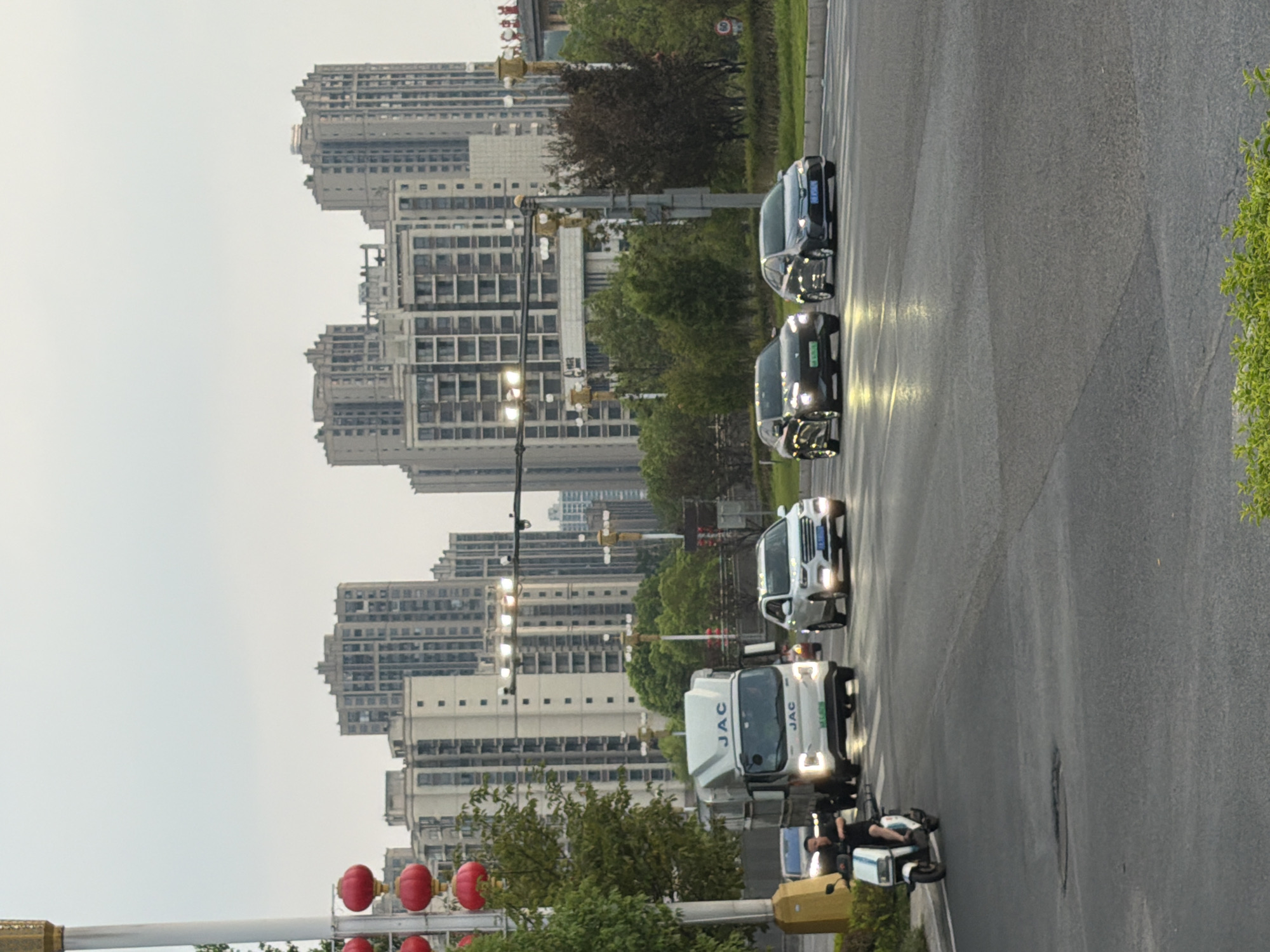 In Jiangjin, China, on an overcast day, a street scene unfolds. A white JAC truck is in the middle lane, with a man on a white scooter in the left lane approaching from behind. To the right of the truck, a white SUV is in the right lane, and further ahead, two dark sedans are in adjacent lanes. The road is wet, reflecting the headlights of the vehicles and traffic lights. In the background, a cluster of tall, modern apartment buildings rises against the grey sky. Red lanterns hang from a pole on the left side of the street, near a lamppost and a small tree. Green foliage is visible along the roadside, and to the far right, a speed limit sign indicating 60 is partially visible.