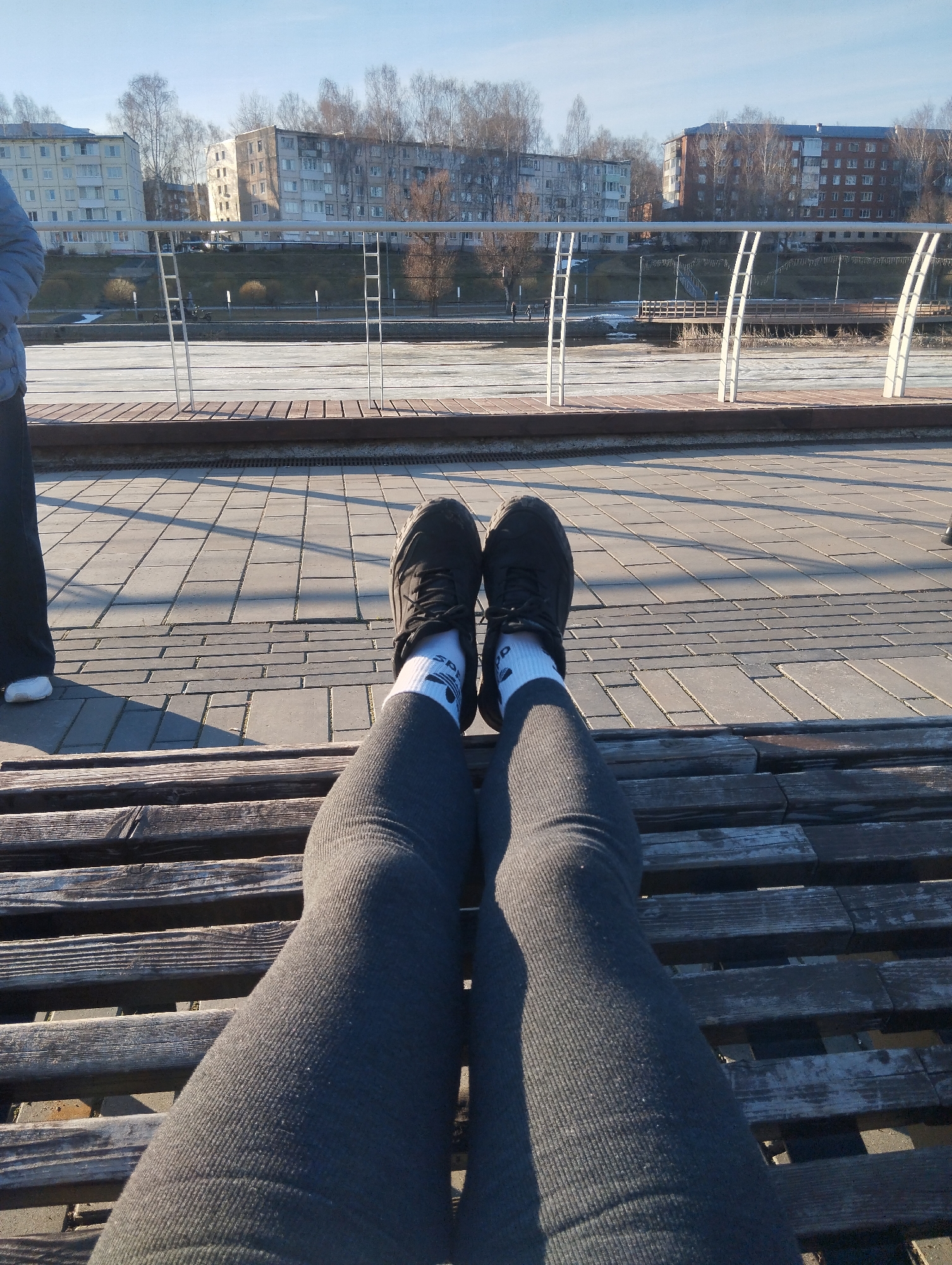 A first-person perspective shot from someone sitting on a wooden bench on a sunny day. The viewer's legs, clad in gray leggings and white socks with black Adidas stripes, are stretched out. Black sneakers are on their feet. The bench is made of weathered, dark-stained wooden slats.
Beyond the bench, there's a paved area with rectangular stone tiles. A low white metal railing runs along the edge of the pavement, separating it from a body of water that appears to be partially frozen. In the distance, there are multi-story residential buildings, typical of Soviet-era architecture. Bare trees are interspersed among the buildings. The sky is clear and blue. Another person is partially visible to the left side of the frame.
The location is Votkinsk, Russia, based on the architectural style of the buildings and the presence of a partially frozen riverfront scene.