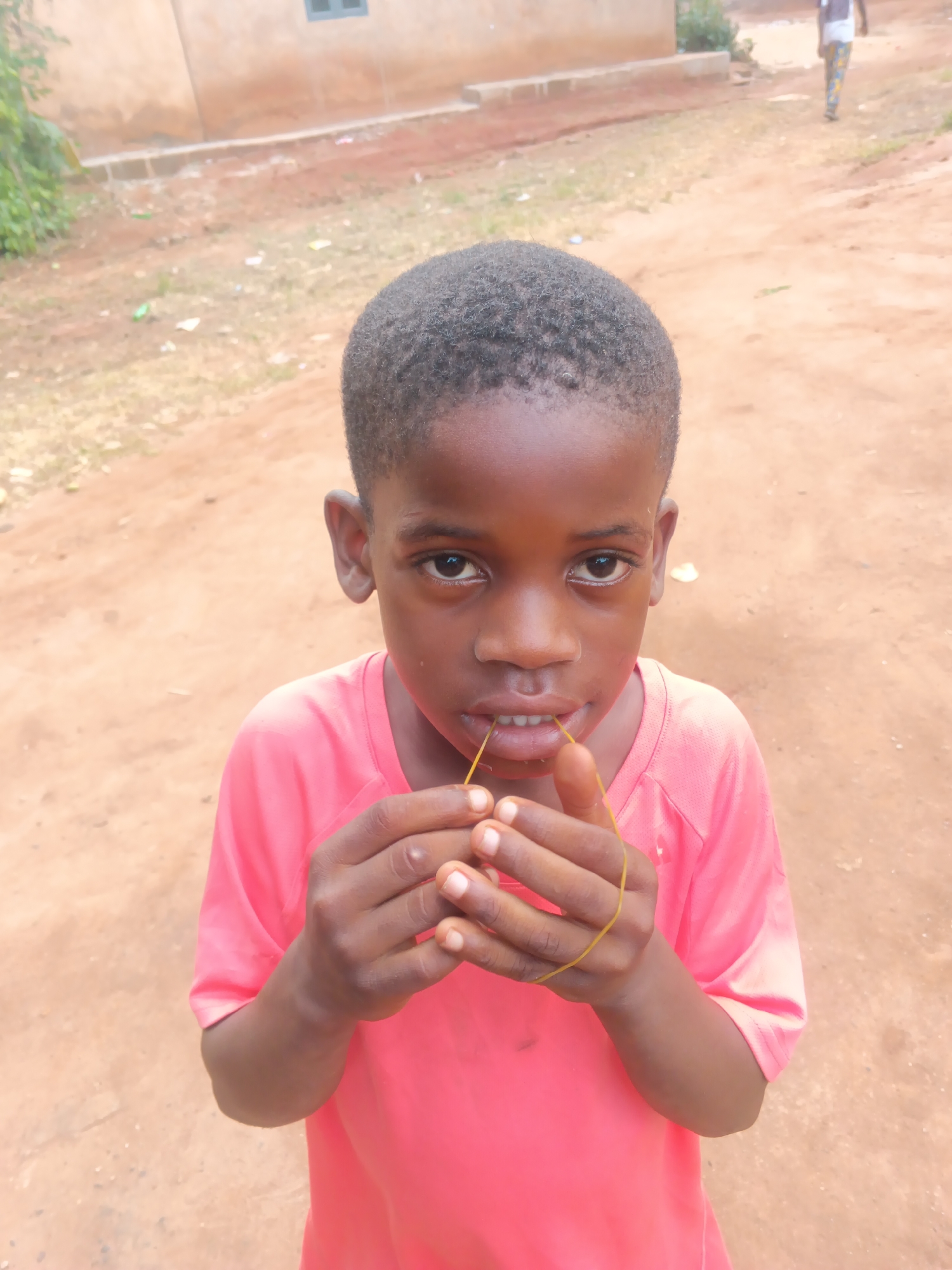 A young boy, likely of African descent, is captured in a close-up, eye-level outdoor portrait. He has short, dark, curly hair and is dressed in a bright coral-pink, short-sleeved t-shirt. The boy looks directly at the camera with a neutral yet focused expression. He is actively engaged with a thin, yellow string, holding it between the fingers of both hands and also gently gripping it with his front teeth.
The setting is an unpaved, reddish-brown dirt ground, characteristic of a rural or semi-urban environment in Eziudo, Nigeria. In the background to the left, a light brown building with a dark window frame is partially visible, flanked by green foliage. Further back on the right, another person in patterned trousers walks away from the camera on a dirt path. The scene is illuminated by clear daylight. There is no visible text in the image.