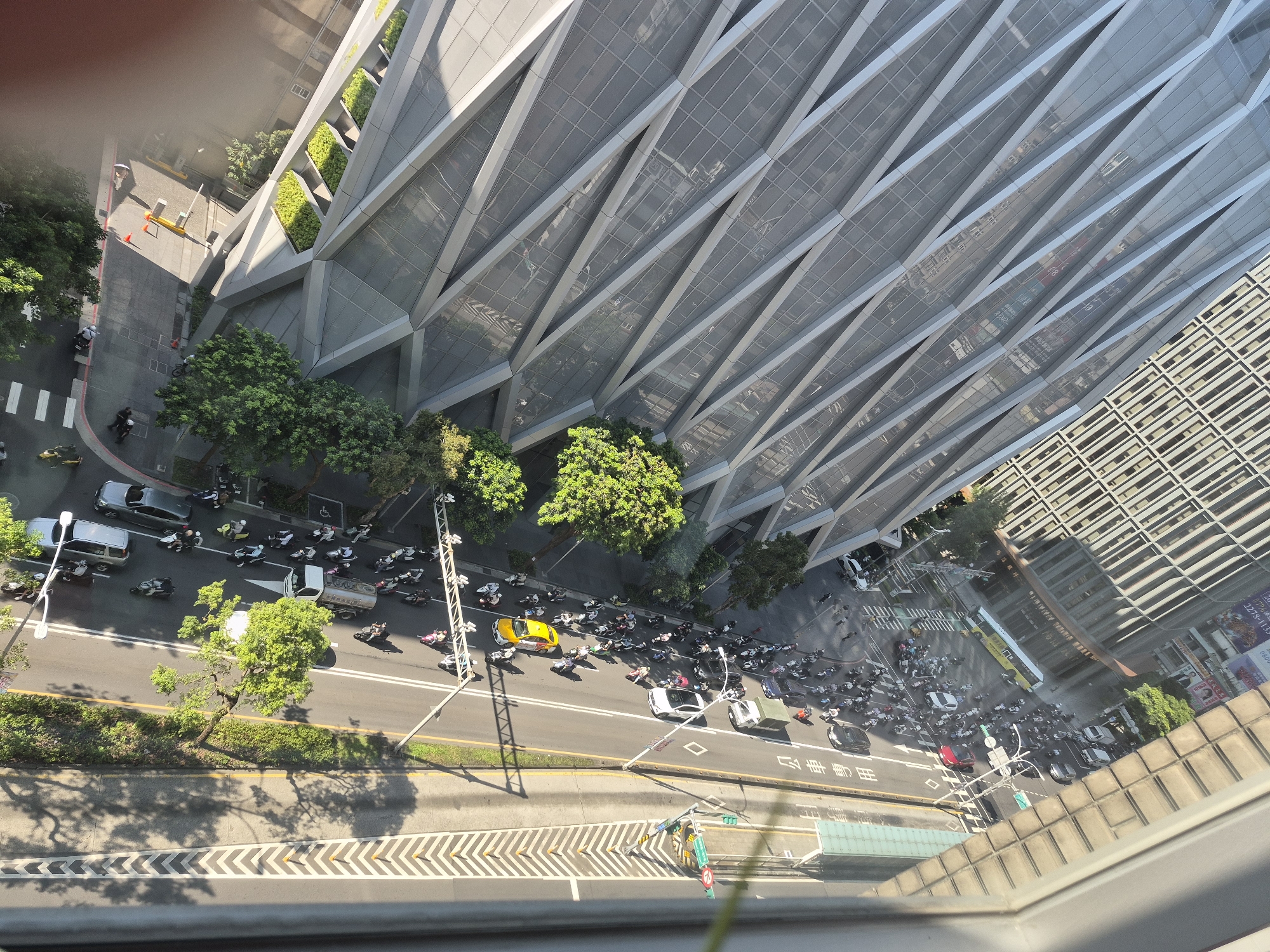 The image, captured from a high vantage point, shows a busy street scene in Taipei, Taiwan. The perspective is a top-down, angled view of a multi-lane road bordered by modern buildings and lush green trees. Numerous motorcycles and scooters are in motion, suggesting a typical urban commute. Several cars, including a silver SUV, a lighter colored sedan, and a truck, are also present on the road. A bright yellow car stands out among the vehicles. Pedestrians are visible on the sidewalks and crosswalks. The architecture of the surrounding buildings is contemporary, with one building featuring a distinctive geometric facade and another displaying a grid-like pattern. The presence of streetlights and traffic signals indicates an active urban environment. The lighting suggests it is daytime, with shadows cast by trees and buildings. The road surface is asphalt, marked with white lines for traffic lanes and pedestrian crossings.