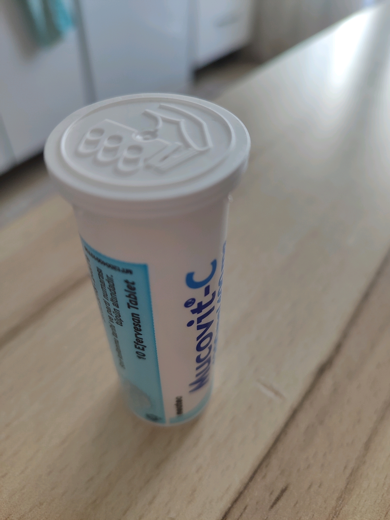 A white plastic tube with a blue label is placed on a light-colored wooden table. The lid of the tube has a raised pattern. The label on the tube is partially visible and contains blue text. The visible text includes "MUCOVIT-C" and "10 Efervesan Tablet". The background is out of focus and appears to be an indoor environment with white cabinets and a light-colored wall. The overall lighting suggests an indoor setting, possibly a kitchen or bathroom, illuminated by natural light. The image has a shallow depth of field, with the tube in sharp focus and the background blurred. There are no people or animals present in the image. The scene is static and does not depict any action or interaction. There is no information in the image that directly indicates the location of Gümüşova, Turkey.