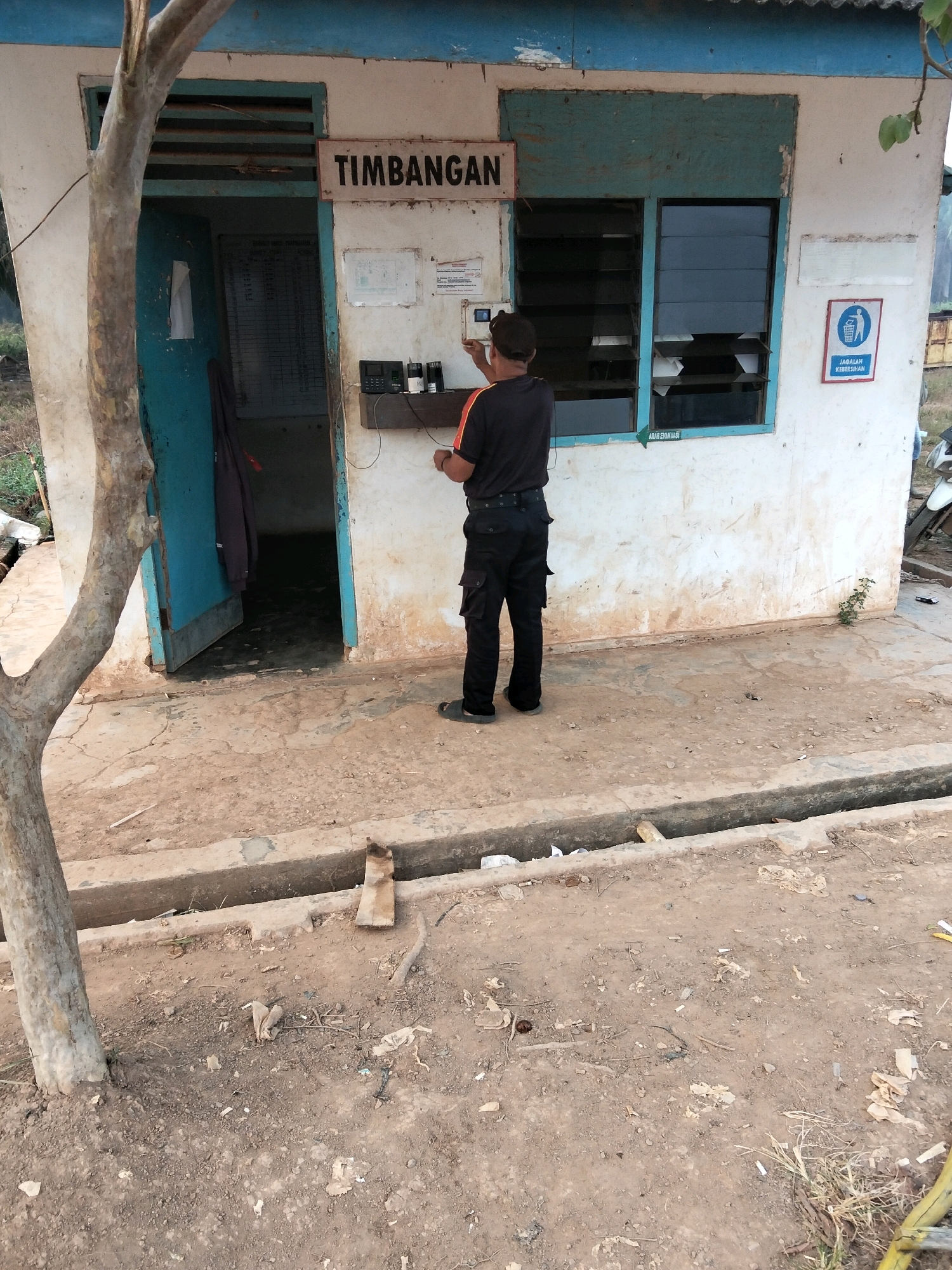 A man in a black shirt and pants is standing outside a small, weathered building with a sign that reads "TIMBANGAN". He is interacting with a small electronic device mounted on the wall next to a door. The building appears to be a weigh station or a small office. The environment is dusty and somewhat neglected, with a drain or ditch running in front of the building. There is a tree in the foreground to the left. The building has windows with louvers and a blue painted frame. A sign with a recycling symbol and text in Indonesian is visible on the right side of the building. The ground is dirt and concrete.