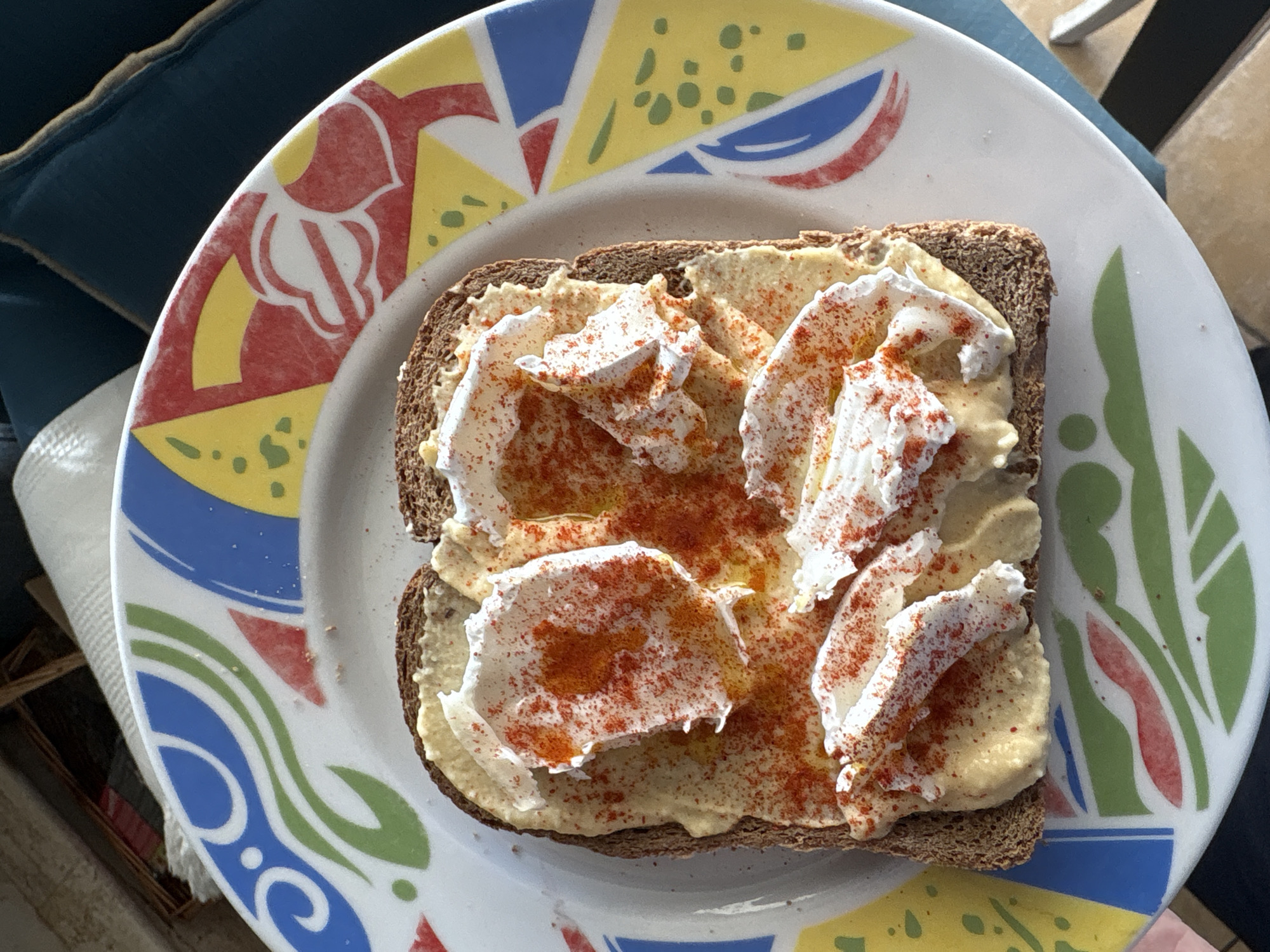 A single slice of whole wheat bread, generously spread with hummus, topped with dollops of what appears to be ricotta cheese, and sprinkled with paprika, sits on a white plate with a multicolored geometric pattern. The plate rests on a surface with potential sunlight. The location is Urbanización Encinares De Alcolea, Spain. No people are visible.