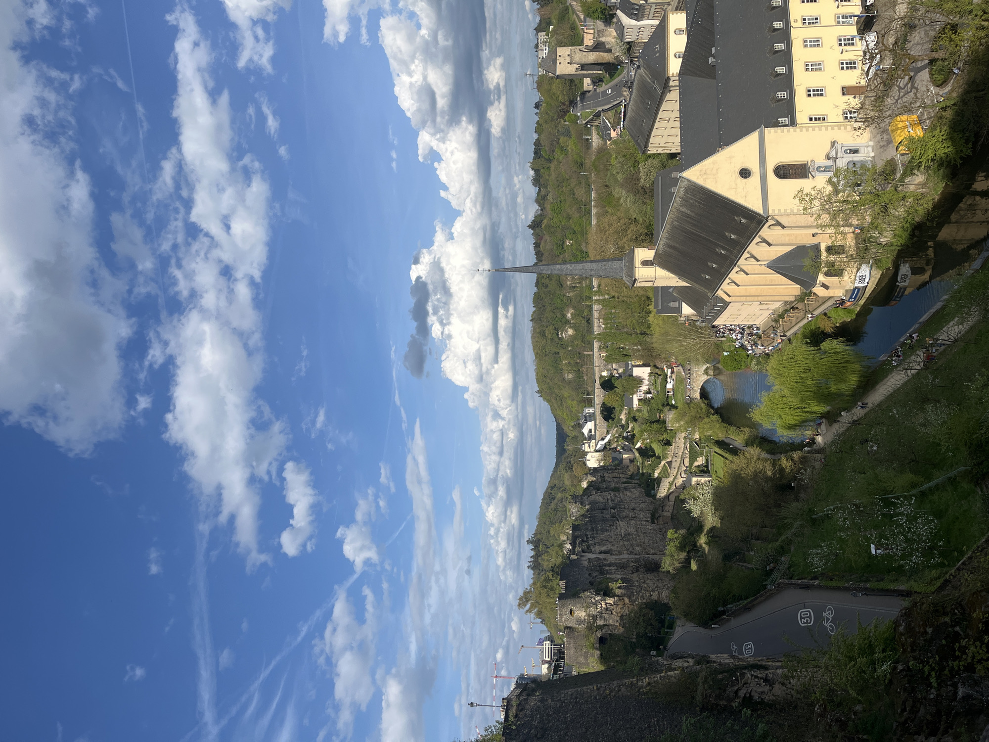 The image is an elevated view of Luxembourg City, Luxembourg, on a sunny day with blue sky and scattered clouds. The scene encompasses historic buildings, a river, and natural rock formations. Prominently featured is the Neumünster Abbey complex, including its church with a steeple. A body of water, likely the Alzette River, borders part of the abbey. People are visible on the banks of the river near the abbey. The landscape includes trees and vegetation. The angle and composition of the photograph suggest it was taken from a high vantage point. A road with bike lanes is visible below the viewpoint.