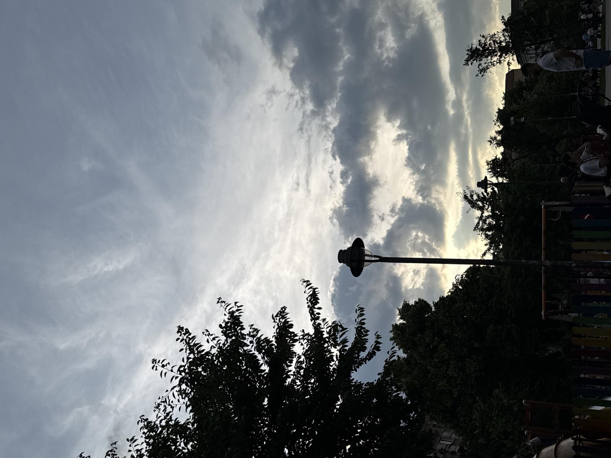 This image captures a park or public green space in the city of Sofia, Bulgaria, during dusk or early evening. The sky is predominantly cloudy, featuring high, wispy cirrus-like clouds in the upper left transitioning to denser, darker grey stratocumulus clouds across the mid and right sections, with lighter, illuminated patches visible near the horizon, suggesting the position of the setting sun.
In the foreground, a multi-colored picket fence, painted in vertical sections of red, orange, yellow, green, and blue, runs across the bottom of the frame, marking the boundary of what appears to be a children's playground. Part of a dark-colored slide structure is partially visible behind the fence on the far left.
A tall, dark lamppost with a bell-shaped lantern top stands slightly left of the image's center, silhouetted against the lighter sky. To its left, a large, densely foliated tree occupies the upper left quadrant and extends downwards, its leaves appearing as dark silhouettes. More dark, mature trees form a dense band across the midground and background. Another similar lamppost is discernible further back on the right side.
Several individuals are present in the scene, mostly in silhouette or with limited detail. On the right side of the midground, a man with short hair, wearing a light-colored t-shirt and light blue shorts, stands with his hands on his hips, facing towards the left. To his left, another person is seated on a low structure or bench, largely obscured. Multiple blurred figures are visible in the background, indicating a populated area. Distant buildings with visible window patterns are faintly silhouetted against the sky in the far background.