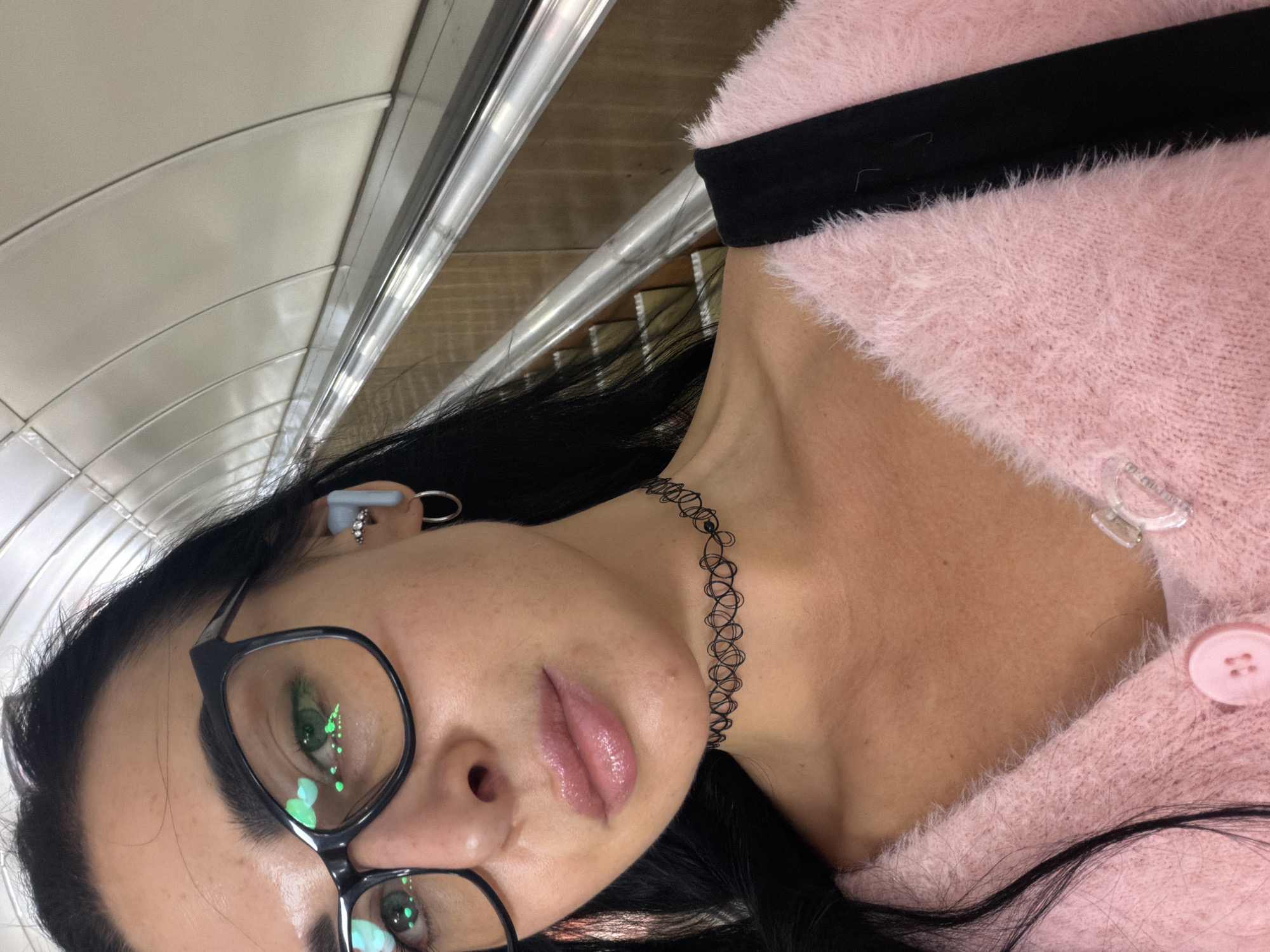 The image depicts a woman with long black hair, wearing black-framed glasses, a black choker necklace, and a pink fuzzy cardigan with black trim. She has a piercing in her left ear and an earbud in her left ear. The background suggests a subway or train station setting, indicated by the curved ceiling, metal bars, and interior of what might be a train car. The woman is likely in Saint Petersburg, Russia, based on the distinctive design of the subway car's interior, which is characteristic of the Saint Petersburg Metro.