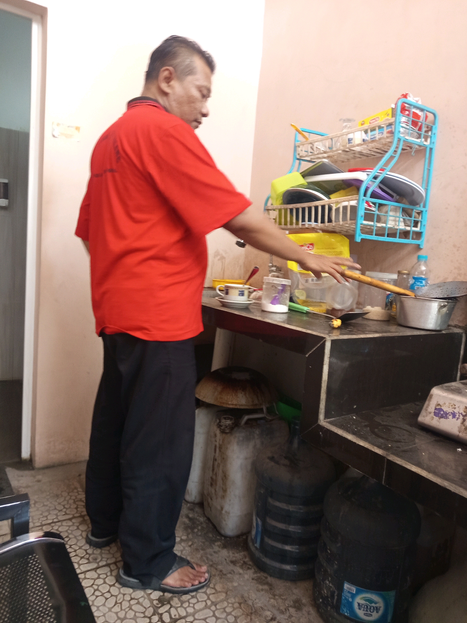 An adult male wearing a red t-shirt and black pants stands in a kitchen area. He is reaching out his right hand towards a wooden utensil on a countertop. The countertop is cluttered with various items, including a metal pot, a plastic container, a cup and saucer, and a bottle of water. A blue dish rack filled with plates and bowls is mounted on the wall behind the countertop. In the background, there are several large water jugs. The scene appears to be a simple, somewhat disorganized kitchen, possibly in a residential or small commercial setting in Malang, Indonesia, given the presence of the "AQUA" branded water jug. The lighting is dim, suggesting indoor lighting.