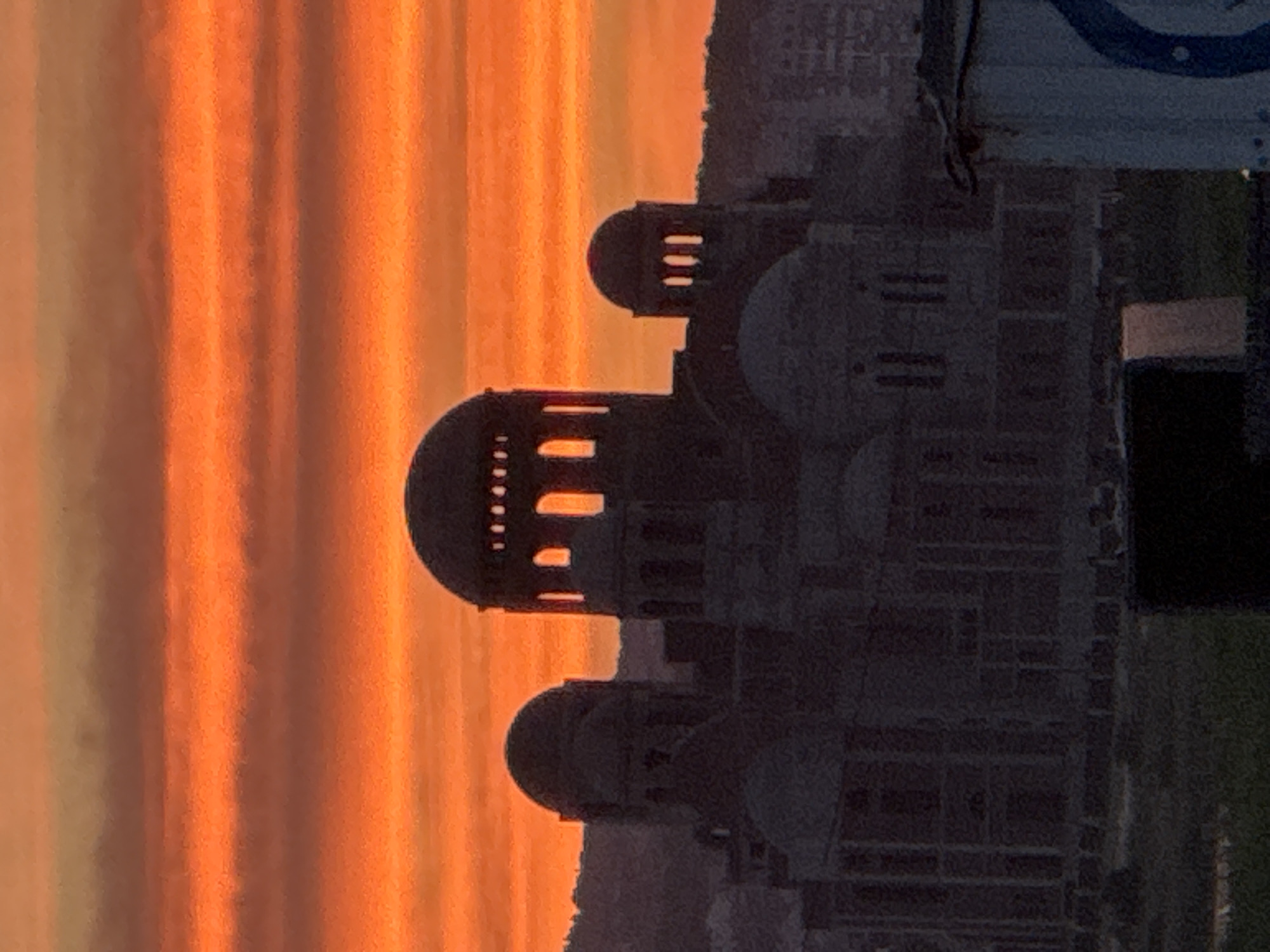 This is a vertical, medium-shot photograph taken at dusk, showcasing the St. Nicholas Naval Cathedral in Novorossiysk, Russia. The sky is a vibrant orange, streaked with horizontal bands of darker red and lighter yellow, indicative of a sunset. The cathedral, a large stone structure with multiple domes and arched windows, is silhouetted against the colorful sky. Some windows glow with an orange light from within. The cathedral appears to be partially under construction or undergoing renovation, as indicated by the unrendered facade in some areas. In the foreground, there is a dark object that is difficult to identify clearly, and to the right, a portion of a white structure with a blue symbol is visible. The overall impression is one of grandeur and solemnity, with the dramatic sunset adding a spiritual or dramatic quality to the scene.