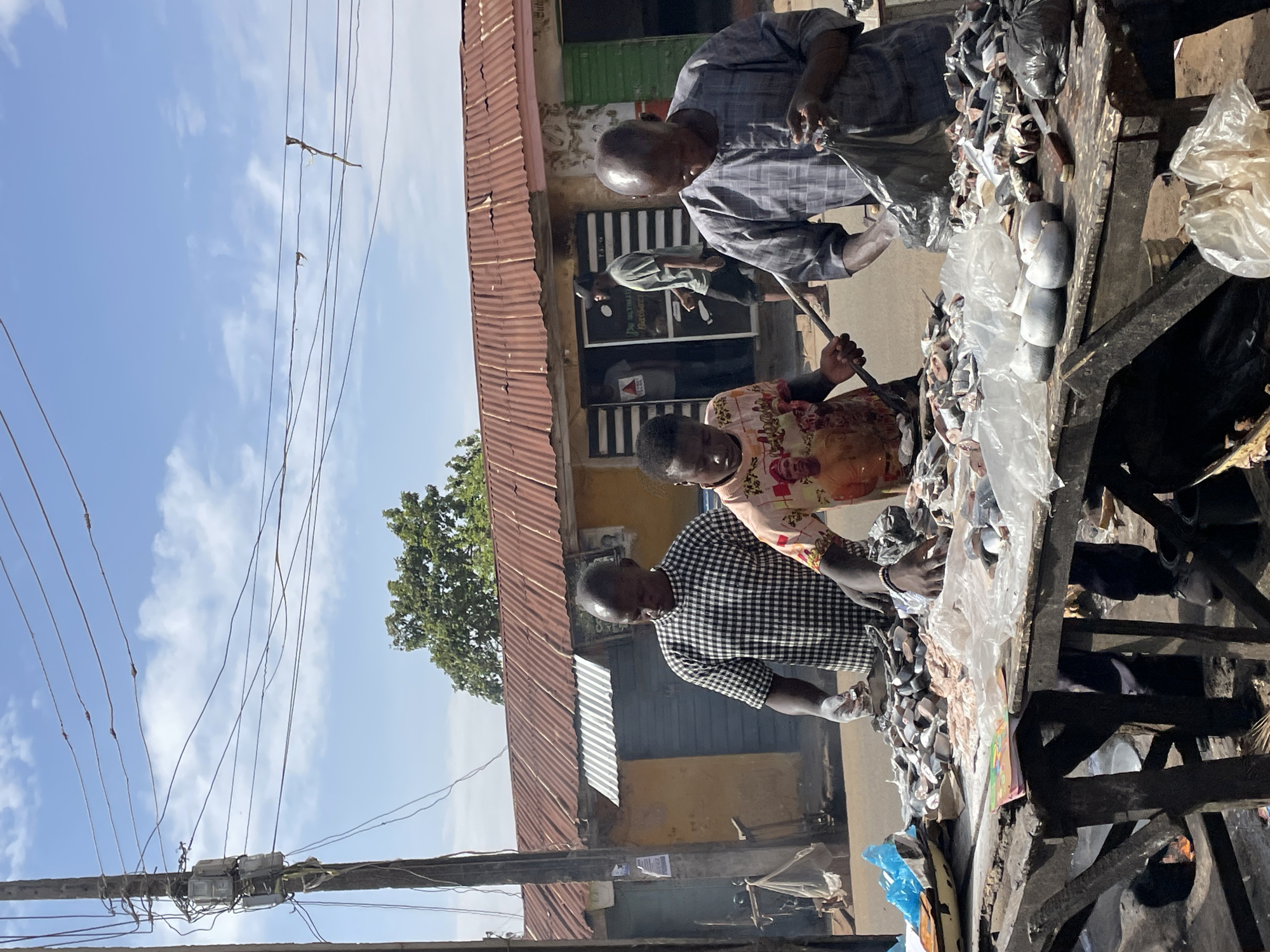Three people are working at a fish market in Jos, Nigeria. They are standing behind a wooden table piled with fish, some of which are in plastic bags. One person is wearing a checkered shirt, another a floral-patterned shirt, and the third a striped shirt. The scene is outdoors, with a building with a corrugated metal roof and utility poles visible in the background. The sky is visible.