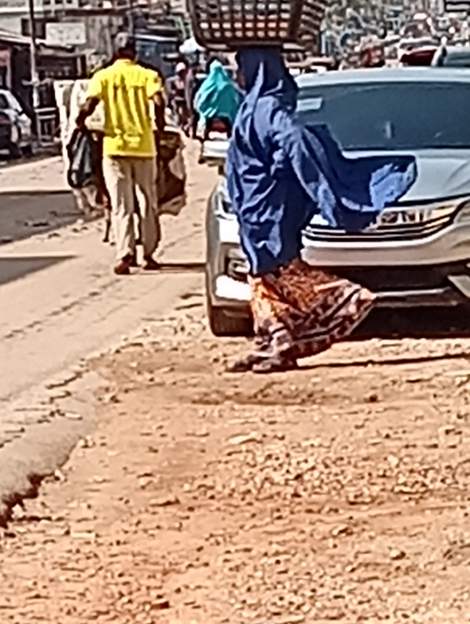The image, apparently taken in the city of Maiburiji, Nigeria, depicts a street scene featuring several individuals and a car. A woman wearing a long blue dress and head covering walks near the front of a white car, carrying a basket on her head. Behind her, a man in a yellow shirt and beige pants carries bags or bundles. Another individual in a blue outfit can be seen further in the background. The street appears unpaved and somewhat uneven. Other vehicles and buildings are visible in the distance.