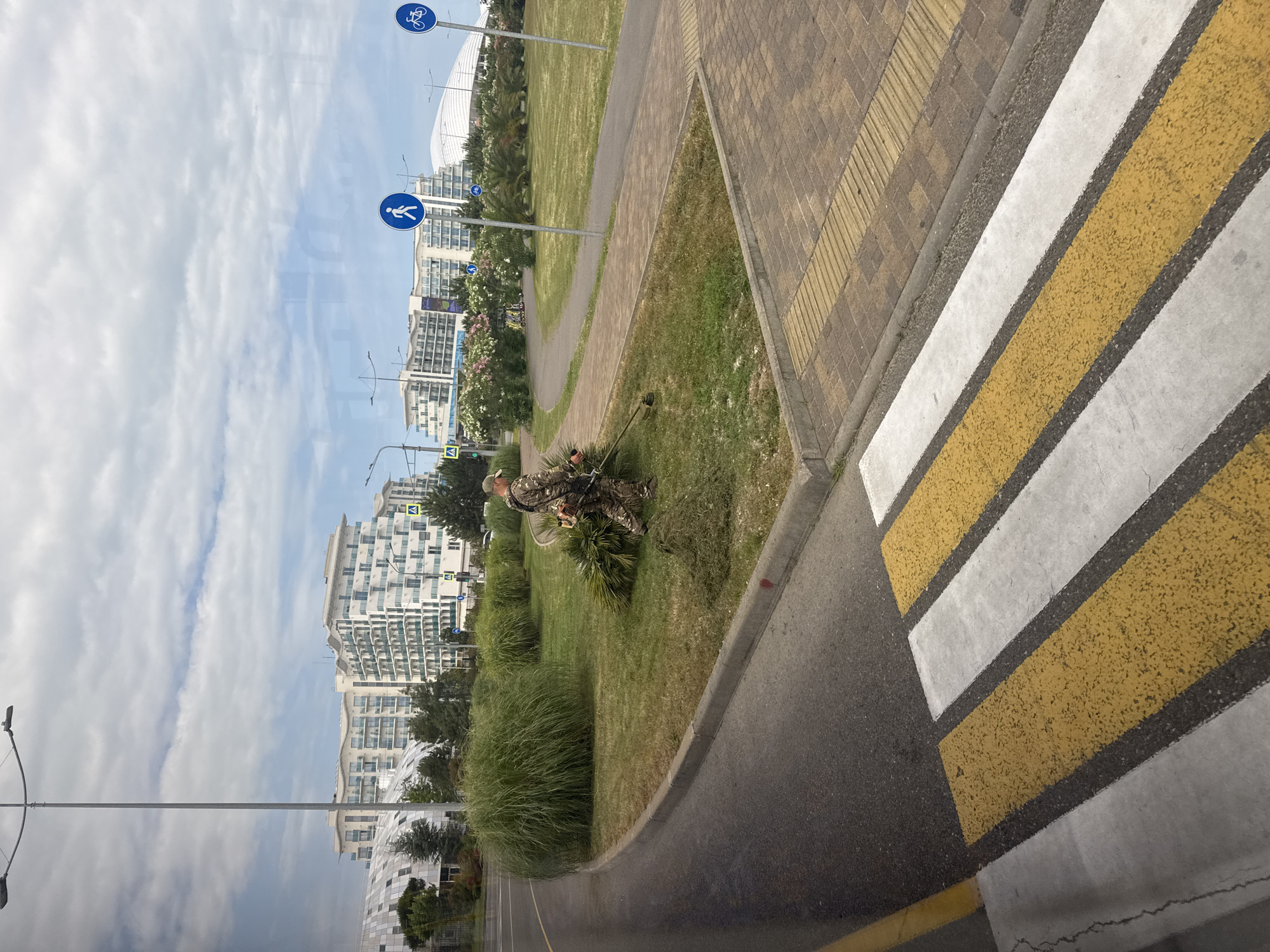 This media file depicts a street scene in Sochi, Russia. The image is oriented vertically. In the foreground, a pedestrian crossing with white and yellow stripes is clearly visible, bordered by a sidewalk paved with yellow and brown bricks and a curb. Adjacent to the sidewalk is a paved road, and then a grassy area. A man wearing camouflage clothing is present, holding a string trimmer, and appears to be engaged in landscaping work, possibly trimming the grass.
In the background, several modern, multi-story buildings with balconies and glass facades are visible, suggesting an urban or resort area. There are also streetlights and some trees and flowering bushes. Blue circular traffic signs with white icons indicating a bicycle path and a pedestrian path are mounted on poles.
The sky is partly cloudy, with a mix of white clouds and blue sky, suggesting daytime. The lighting is bright, consistent with daylight hours. The overall impression is of a well-maintained urban environment. No visible text is clearly legible within the image.