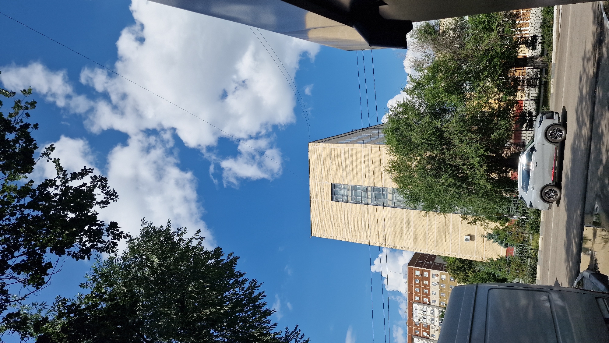 The image shows a street scene in Kostanay, Kazakhstan, on a bright, sunny day with a clear blue sky dotted with fluffy white clouds. In the mid-ground, a distinctive, modern building with a light yellow brick facade and a tall, narrow, multi-paned window dominates the frame. This building has an angular, almost trapezoidal shape. To the left of this building, partially obscured by trees, is a red-brick residential building with multiple floors and windows.
In the foreground, a light gray car is driving on the asphalt road. The car has a red stripe along its lower body. To the left, a portion of a large, light gray van or truck is visible, parked on the side of the road. The road surface appears wet in places, suggesting recent rain or sprinklers, with reflections visible. Trees with lush green foliage line the street, and a dark metal fence is visible behind the car. Overhead, tree branches with dark leaves frame the upper portion of the image. Power lines are also visible stretching across the sky.