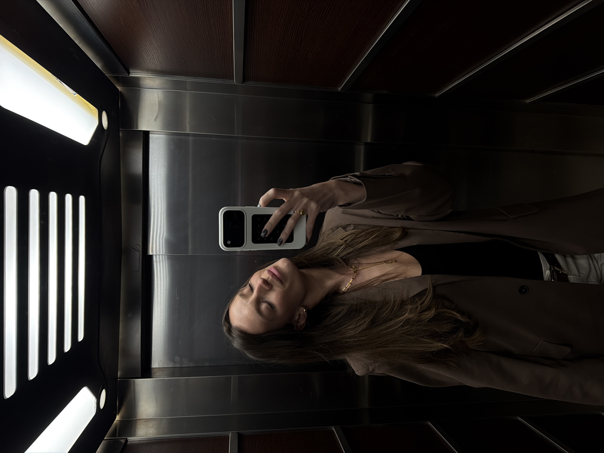 The image depicts a woman's selfie taken inside an elevator, presented with a 90-degree clockwise rotation. The woman, positioned centrally, has long dark brown hair and a relaxed expression with closed eyes, her head gently tilted. She is wearing a dark top, a gold chain necklace, a brown blazer, and a gold ring on her finger. Her nails are painted dark. She holds a white smartphone, featuring dark camera lenses, to capture her reflection in the elevator's polished metallic wall.
The elevator interior is characterized by highly reflective silver-toned panels on the walls. The ceiling consists of a dark, possibly wooden, textured material. On the left side, bright rectangular light fixtures are prominent, some integrated into a black panel with vertical slots, providing strong artificial illumination. This lighting creates significant reflections and plays of shadow throughout the enclosed space. The setting is consistent with an elevator in a modern building, and the provided context places this location in Budva, Montenegro. There is no discernible text visible in the frame. The overall impression is one of a quiet, introspective moment.