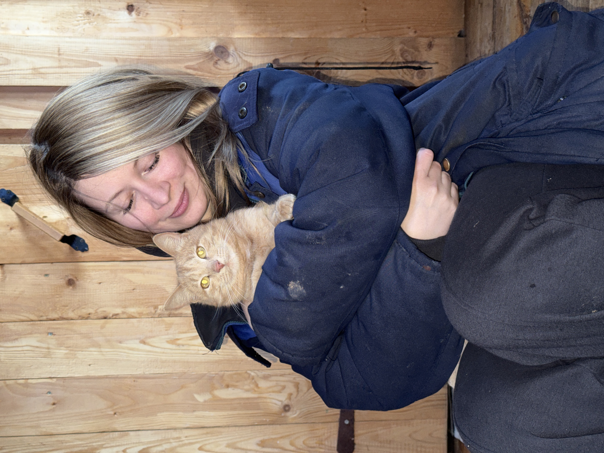 A woman with blonde hair holds an orange tabby cat. The woman wears a dark blue, heavy jacket with multiple buttons and is seated. The background consists of unfinished wood paneling. A tool with a black handle rests on the wood paneling near the woman's head. The location is Вассята, Russia.