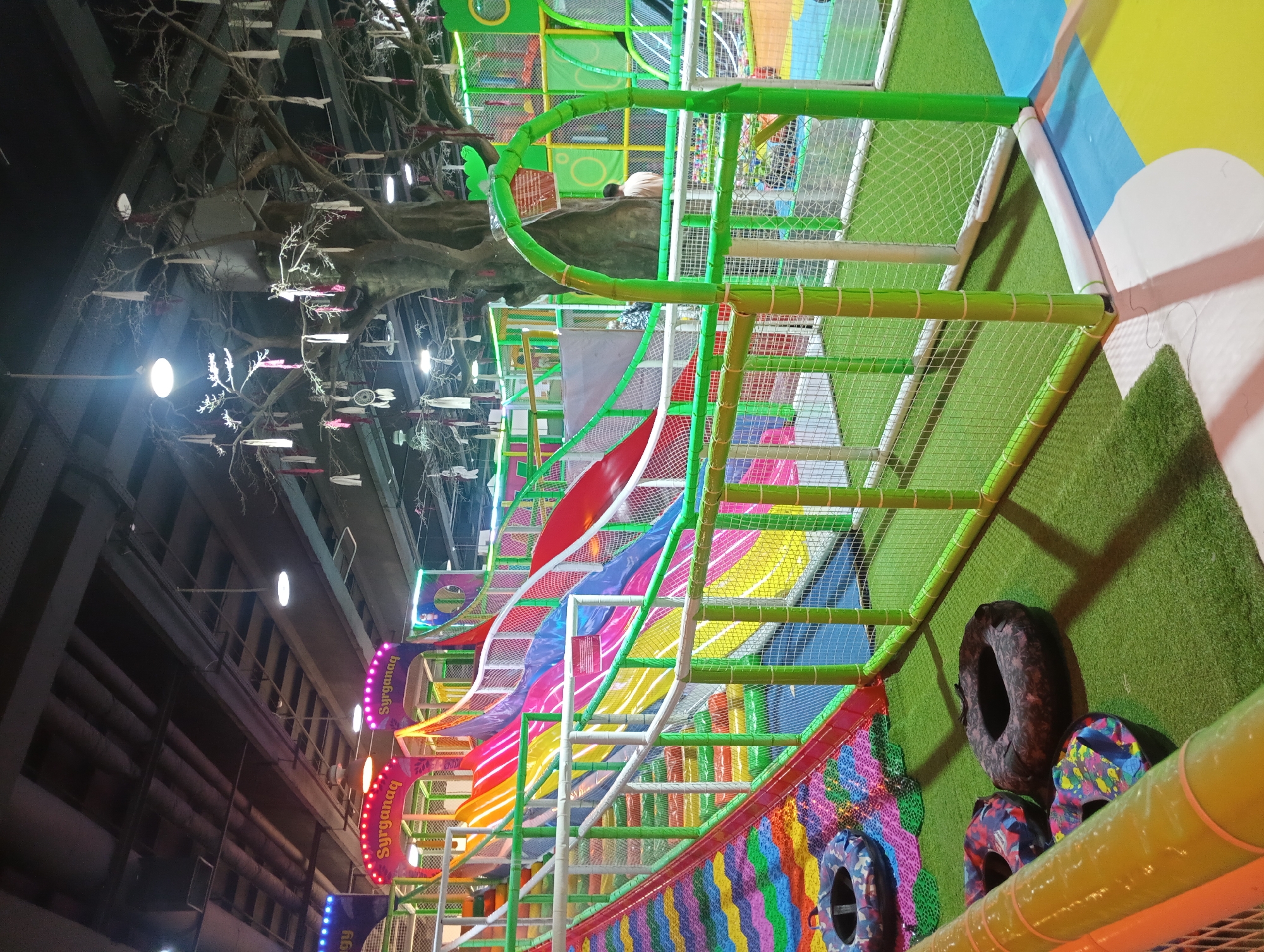 The image depicts an indoor playground, likely located in Almaty, Kazakhstan, as suggested by the signage "Syryganaq" which appears to be a Kazakh word. The playground features a large, colorful slide complex with multiple rainbow-colored slides descending from a central structure. The slides are made of a smooth, glossy material, and are bordered by white and green safety netting. A large, artificial tree with bare branches hangs from the ceiling, adorned with white and red ribbons, creating a whimsical ambiance. In the foreground, several inflatable rings with camouflage patterns are scattered on a green, turf-like flooring. The environment is brightly lit with overhead lights and illuminated signs. In the background, two individuals are visible, one standing near the artificial tree and another partially obscured by the playground structure. The overall scene is one of an active and playful indoor entertainment space designed for children.