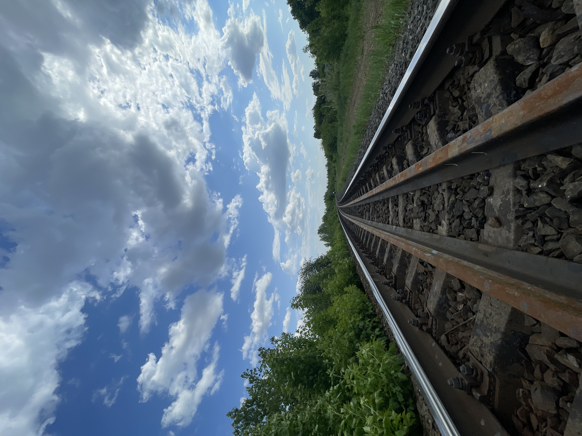 The image shows a section of railway tracks stretching into the distance under a partly cloudy sky. The tracks are made of steel rails and are supported by concrete sleepers embedded in gravel. Vegetation, including grass and trees, flanks the tracks on both sides. The sky is blue with scattered white clouds. The angle of the shot suggests a low vantage point, looking up along the length of the tracks. There is no information in the provided image which proves that it is located in Izhevsk, Russia.
