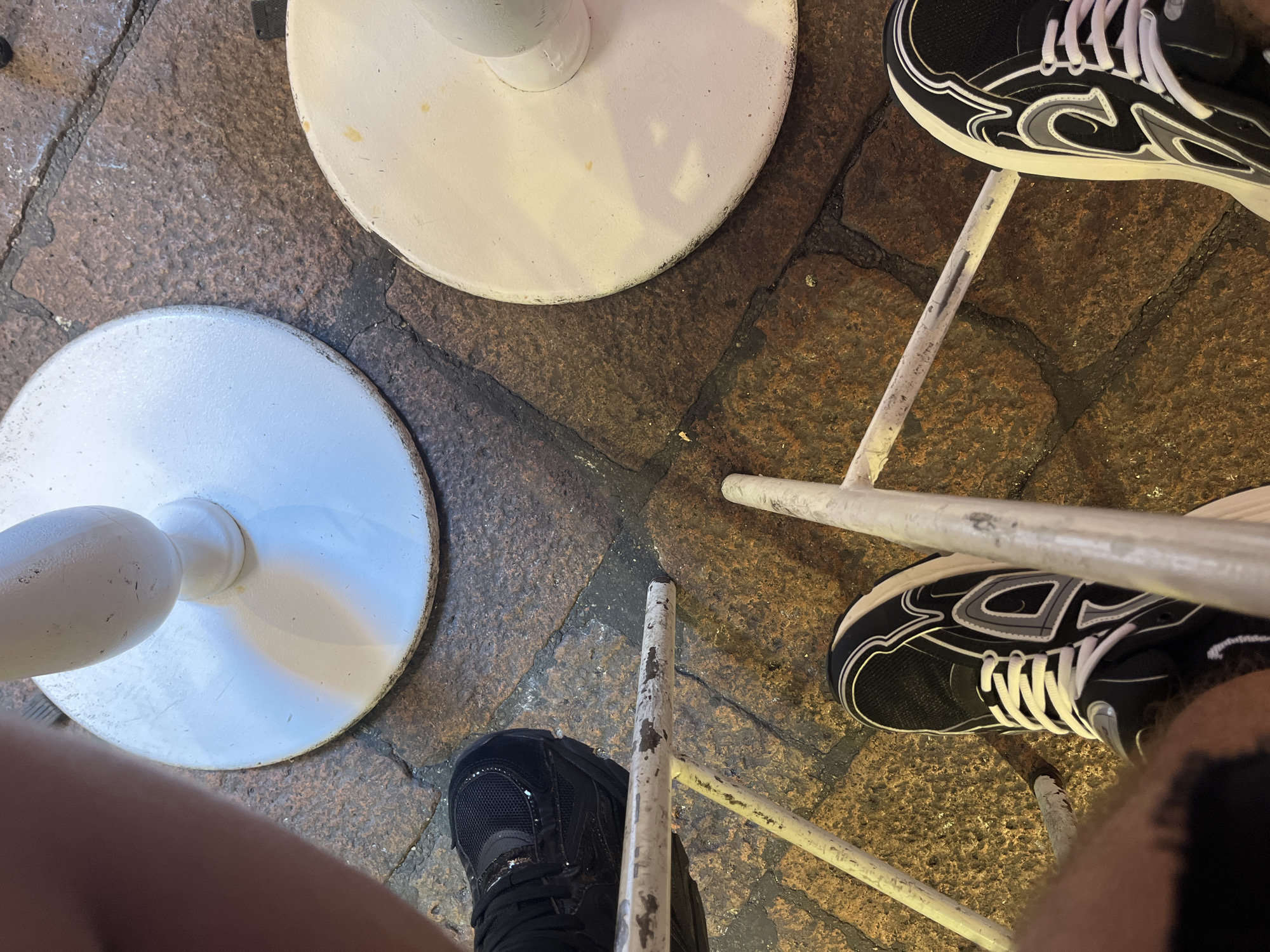 The image shows a close-up, overhead view of two pairs of legs and shoes positioned around a metal chair. One person is wearing black sneakers with white laces and white detailing. The second person is wearing a black shoe. The legs are positioned around two round, white metal bases of the chair's legs. The floor consists of uneven, stone paving. The location is Bologna, Italy, based on the pavement style and the nature of the outdoor setting.