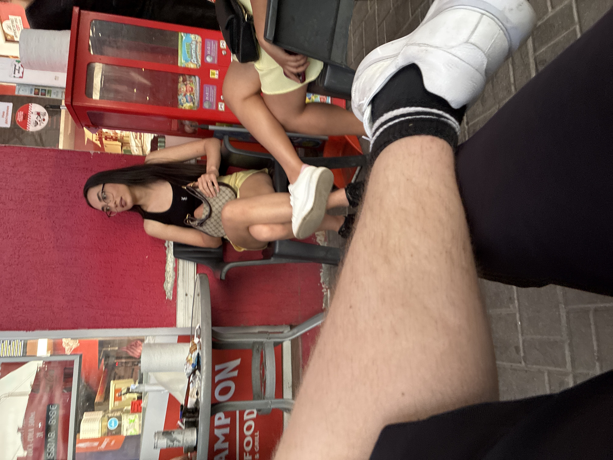 The image depicts a street scene in Burgas, Bulgaria, showing two young women seated at an outdoor table, with a person’s leg and foot prominently in the foreground. The woman on the left is wearing a black top and yellow shorts, holding a patterned bag. The other woman is wearing a yellow dress. There's a red vending machine nearby. The person in the foreground is wearing black pants and a white sneaker with black socks. A table with a food advertisement is visible on the left.
