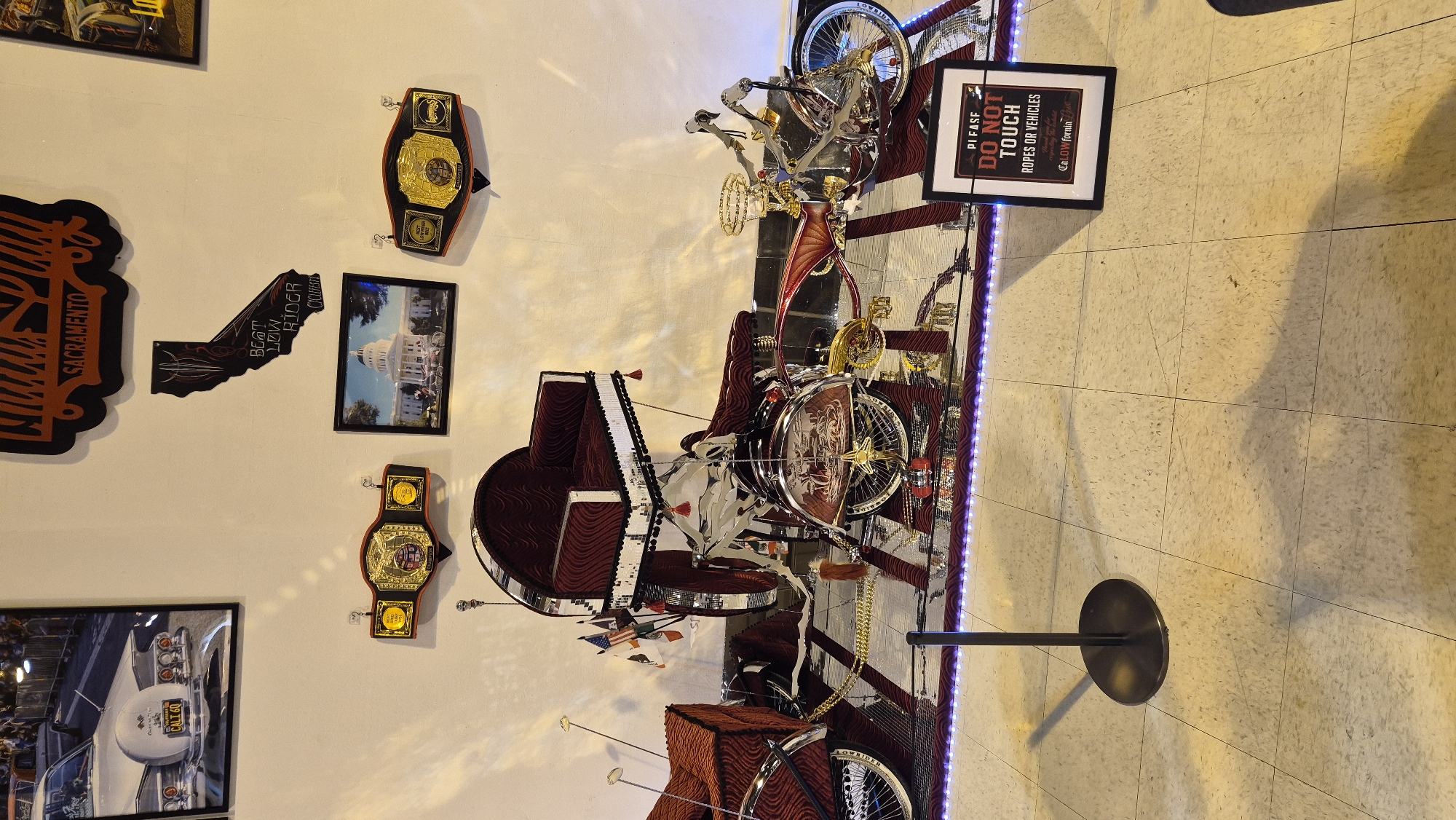 The image captures a display of a custom-built, low-rider bicycle in Sacramento, California. The bicycle is highly ornate, featuring extensive chrome plating, red velvet upholstery on its seats, and elaborate gold accents. It has a distinct, elongated frame and appears to be designed for a single rider, with a prominent seat elevated at the rear. The bicycle is displayed on a mirrored surface that reflects its detailed craftsmanship.
Hanging on the wall behind the bicycle are several items related to car shows and local pride. A large, framed black and orange sign with "SACRAMENTO" written in a stylized font is at the top. Below that, a black silhouette of the state of California contains the text "BEST LOW RIDER". Two championship belts, likely awards from car shows, are mounted on the wall on either side of a framed photograph of the California State Capitol building, which shows a white carriage in front of it. To the left of the display, a framed photograph shows the rear of a white classic car with a California license plate reading "CALT 60".
In the foreground, a black stanchion with a flat circular base stands to the left of the bicycle. To the right, a framed sign with the text "PLEASE DO NOT TOUCH ROPES OR VEHICLES" is placed on the floor. The floor itself is tiled in a light beige color with a subtle pattern. Purple LED lights are visible along the edge of the display platform, illuminating the area around the bicycle. The overall scene suggests an indoor exhibition or museum dedicated to low-rider culture, specifically highlighting Sacramento's connection to this automotive art form.