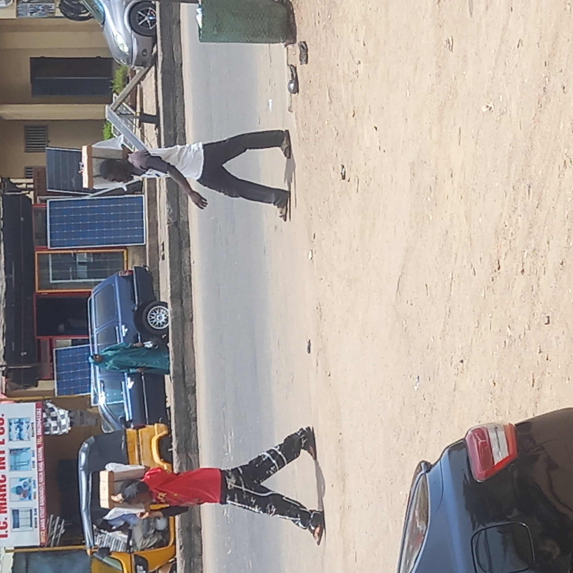 The scene is a street in Dadi Riba, Nigeria. Two men carry items on their heads, one carrying a box on his head, and the other seemingly carrying solar panels. The background reveals a building with solar panels for sale. A parked car and a yellow auto-rickshaw are also visible. The overall environment suggests a commercial area with street vendors or shops related to solar energy.