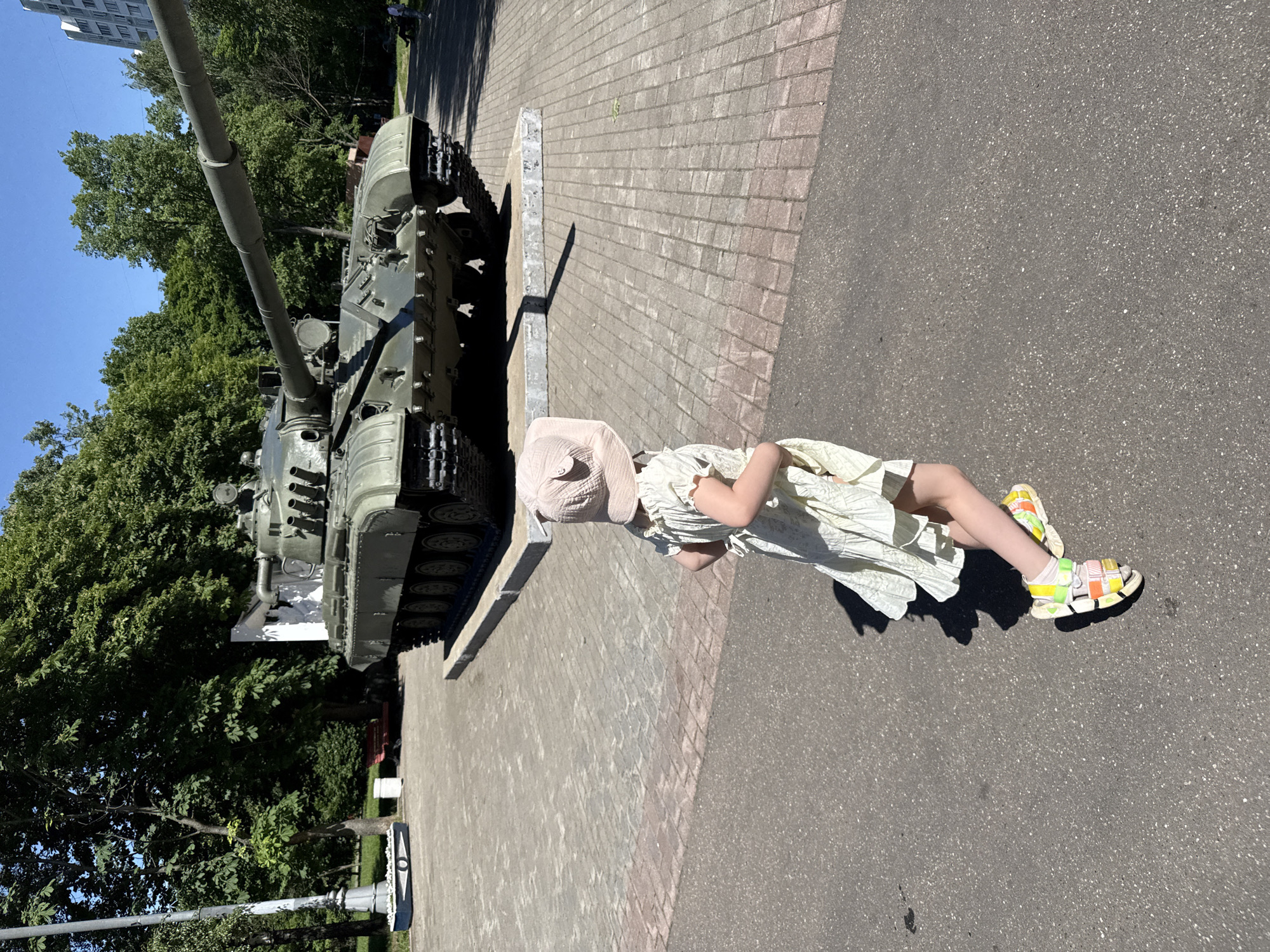 The image depicts an outdoor, sunlit scene at the **Central Armed Forces Museum (Центральный музей Вооружённых Сил)**, located at Sovetskoi Armii St., 2, Moscow, Russia. The foreground and mid-ground consist of paved pathways, featuring asphalt in the immediate foreground transitioning to light-grey rectangular concrete pavers further back, separated by a horizontal line of red-toned pavers.
A young child, observed from behind, occupies the lower central-right portion of the frame. The child wears a light-colored, possibly off-white or pale yellow, sleeveless dress and a light-pink or beige bucket hat with subtle ear-like protrusions. White socks and multi-colored sandals (yellow, orange, pink, green, white straps) adorn the child's feet. The child is in motion, stepping forward with the right leg slightly raised, facing towards the central exhibit. A distinct shadow of the child extends onto the pavement to the right, indicating bright sunlight from a high angle.
The central subject is a large, olive-green main battle tank, identified as a T-72 variant by its six large road wheels and the configuration of its turret and 125mm smoothbore gun. The tank is mounted on a low, grey concrete plinth, positioned in the mid-ground. The tank's long barrel extends diagonally towards the upper-left of the frame. To the left of the tank, a tall, grey street lamp with an ornate base and a blue, boat-shaped flower bed filled with white blossoms are visible. Behind the tank's turret, on the viewer's left, a segment of a white wall or structure is present. Dense green foliage from numerous deciduous trees forms the background, encompassing a faint red park bench on the left. In the far-right background, partially obscured by trees, a tall, light-colored, multi-story residential building with visible windows or balconies rises above the treeline.
The scene is brightly illuminated by direct sunlight, creating sharp shadows and suggesting clear weather. The overall environment is a public park or museum exhibition area.