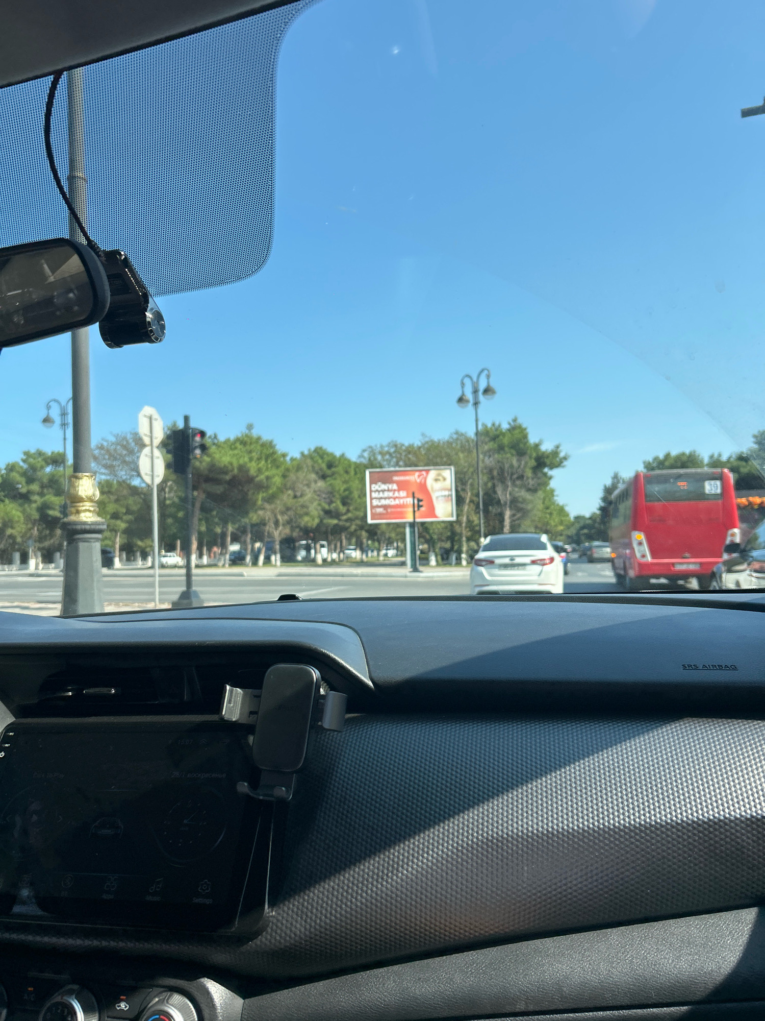 The image is taken from the interior of a car, looking out through the windshield. The car appears to be in motion or stopped in traffic on a sunny day. In the foreground, the dashboard is visible, with a phone mount attached to it holding a smartphone. The windshield has a dotted pattern, likely from a protective film or tint.
Outside the car, a street scene unfolds. On the left, a tall lamppost with a decorative golden base stands next to a traffic light which is red. Further down the street, there are trees lining the roadside and a billboard. A white car is visible on the road, and to its right, a red bus with the number 39 on its rear is also in view. Several other vehicles are present on the road in the distance. The sky is clear and blue. Based on the context of the billboard which appears to have text in Azerbaijani and the presence of a prominent red bus (common in Baku and Sumgayit public transport), the location is likely the city of Sumgayit, Azerbaijan.