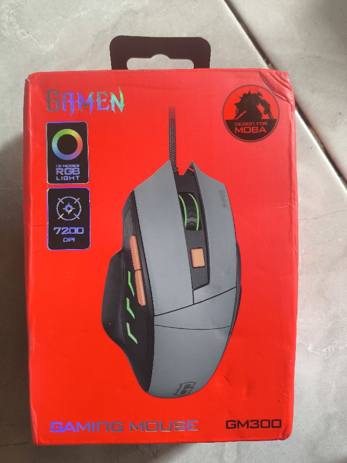 A red box displays a GAMEN GM300 gaming mouse. The mouse is grey with black accents and orange details. The box features a "DESIGN FOR MOBA" logo and boasts "1D MODE RGB LIGHT" and "7200 DPI" capabilities. "GAMING MOUSE" is printed at the bottom in iridescent lettering. A black hanging tab extends from the top of the box.