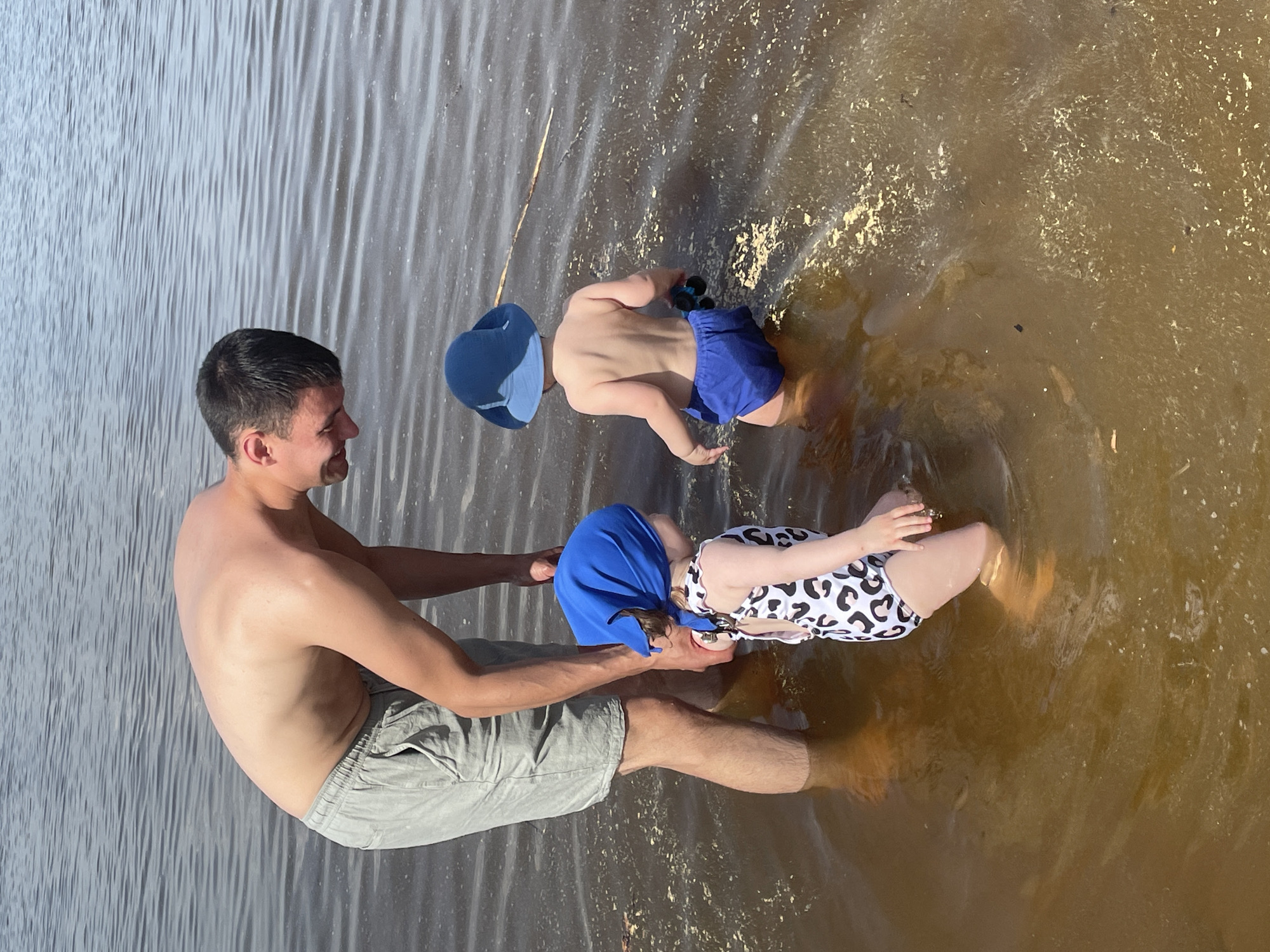 A man, two children, and a body of water are visible in the image. The man is shirtless and wearing gray shorts, holding a young child wearing a blue swim cap and a white and black swimsuit. The child is partially submerged in the water. Another child wearing a blue swim cap and blue swim trunks is also in the water, with their back to the viewer. The water appears to be murky or brownish, and a stick or branch is partially submerged near the first child. The water is still with gentle ripples. It's impossible to verify the location without further information.