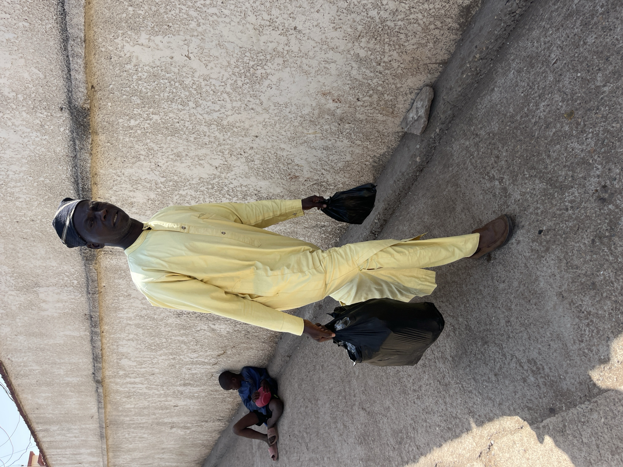 The image shows an adult man dressed in a light yellow tunic and trousers, with a black cap, holding two black plastic bags. He is standing on a concrete surface next to a light-colored wall. A younger person is sitting on the ground near the man, wearing a blue shirt and dark shorts, and holding a red object. The background includes a rough, light-colored wall and a portion of the sky. The location is within the city of Jos, Nigeria, inferred from the context of the image and the typical clothing worn in that region.