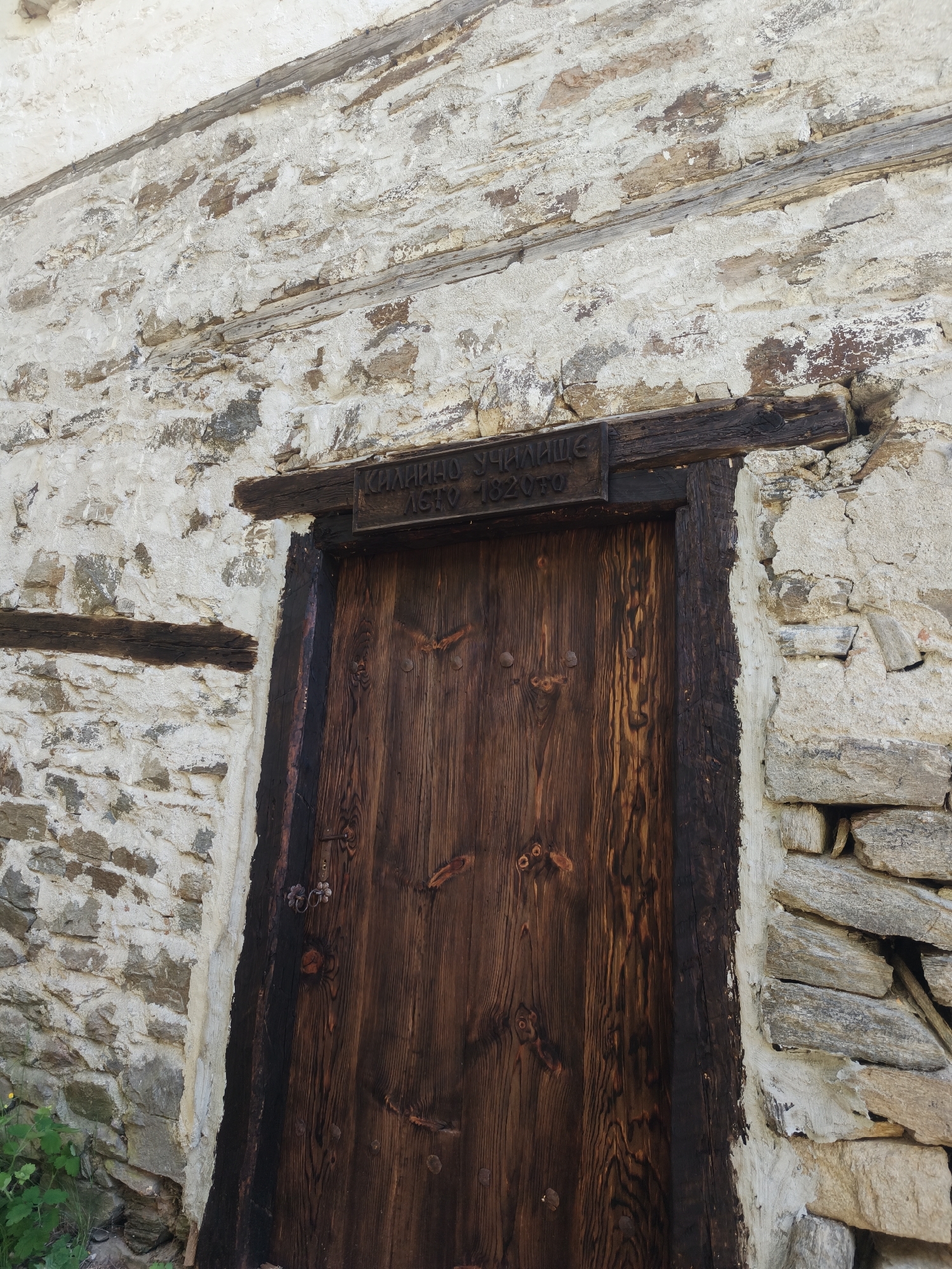 A wooden door is set in a stone wall in Kovachevitsa, Bulgaria. The door is made of dark wood with a vertical grain pattern. A dark wooden frame surrounds the door, and a wooden lintel with an inscription in Cyrillic script sits above the door. The inscription includes the word "УЧИЛИЩЕ" (School) and the years "1870-1970". The wall is constructed of irregularly shaped stones, mostly light in color, and is covered in some areas with what appears to be light-colored plaster or mortar. Green foliage is visible at the bottom left of the image.