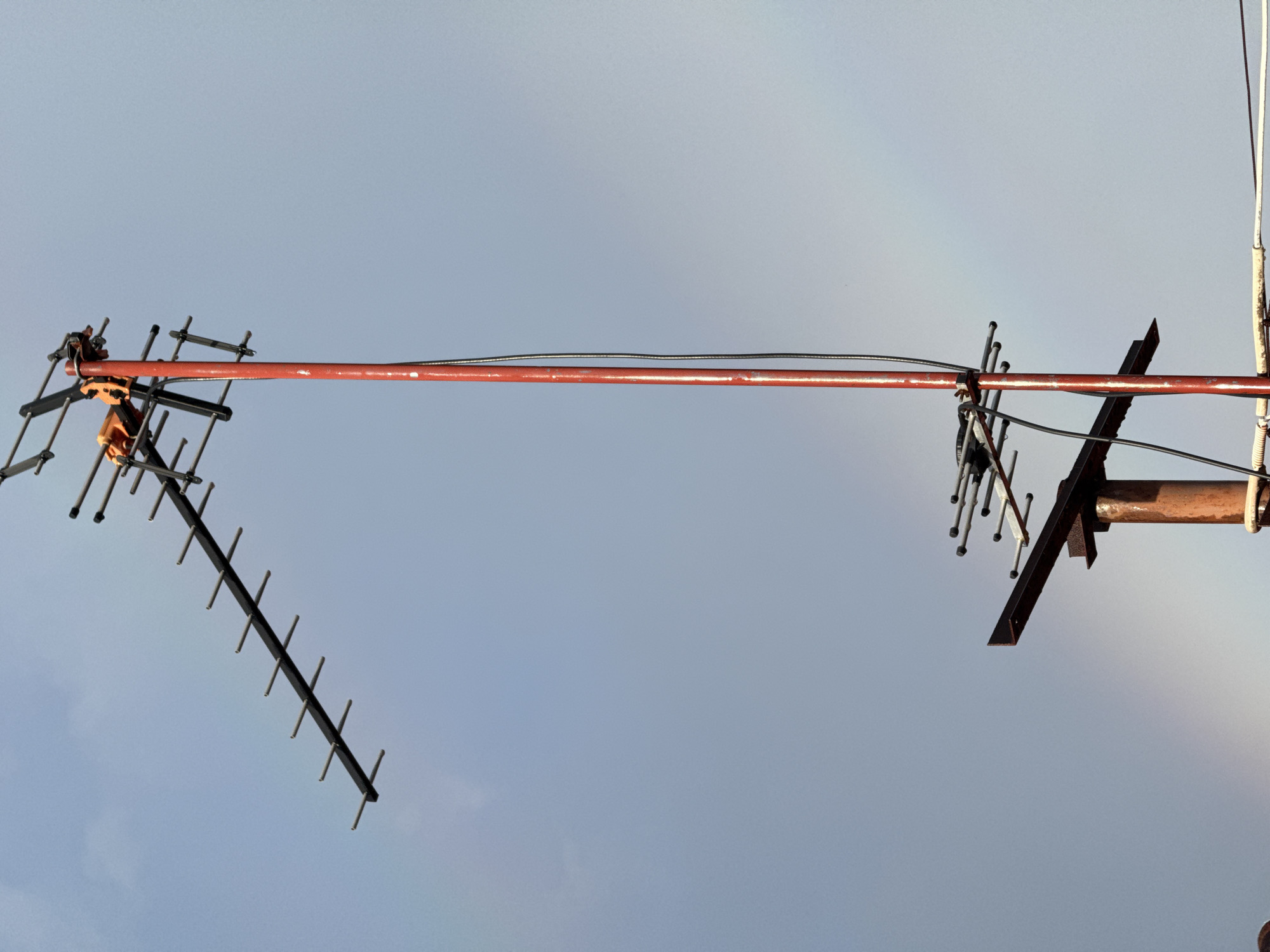 The image shows an outdoor scene with a clear sky, likely taken in the city of Wichit, Thailand. The main subject is a tall, red-painted antenna mast with two antennas attached. The upper antenna is larger and oriented horizontally, while the lower antenna is smaller and also appears horizontal, mounted on a rusty metal support structure. Wires are connected to the antennas and run down the mast. A faint rainbow is visible in the background, suggesting recent rain or atmospheric conditions. The overall scene is a mix of man-made structures against a natural, albeit overcast, sky.