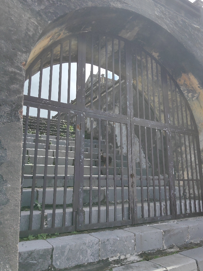 An upward-angled view captures a weathered, dark gray stone archway with a heavy, double-panel metal gate, closed and secured by a padlock, in Phú Xuân Ward, Vietnam. The gate, exhibiting significant paint chipping and rust, features vertical bars that partially obscure a long, ascending staircase beyond. The staircase, constructed of light gray stone or concrete, shows minor green vegetation growing between steps. Approximately halfway up the stairs, a small, dark-colored canine animal is visible, facing the viewer. Further in the background, atop the staircase, a multi-tiered building with traditional East Asian architectural elements and weathered facades is visible against a bright, overcast sky. The overall scene is illuminated by natural daylight.