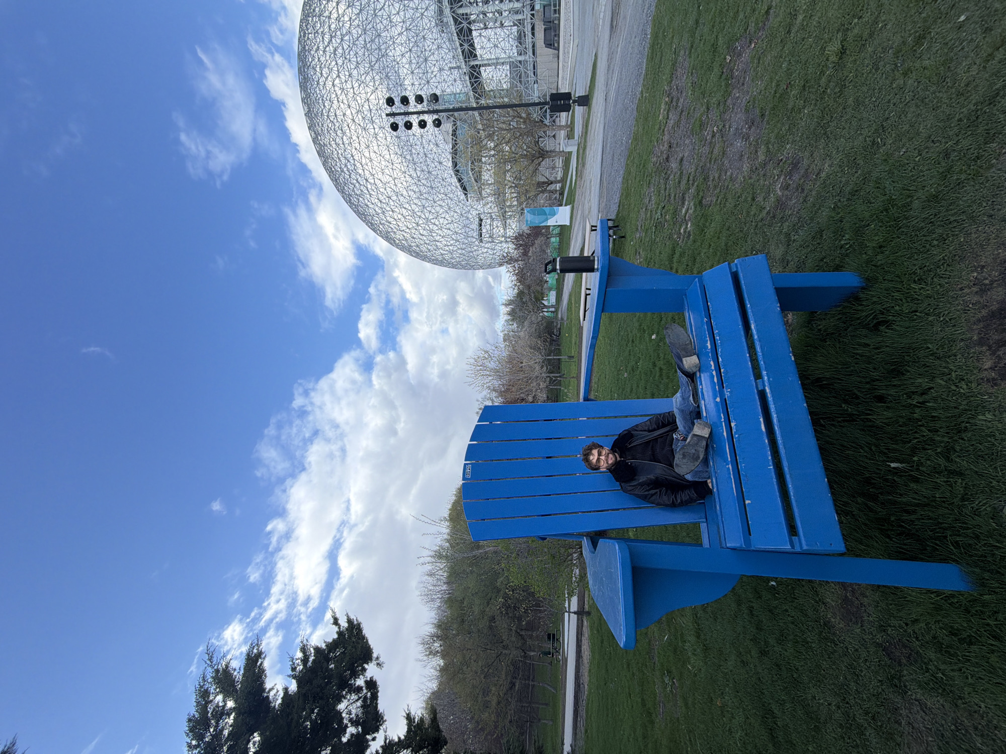 The image depicts a man sitting in an oversized blue Adirondack chair. He is wearing glasses, a black jacket, and blue jeans. The chair is placed on a grassy area. In the background is the Montreal Biosphere, a large geodesic dome structure. The sky is blue with scattered clouds. The location is in Montreal, Canada, near the Biosphere.