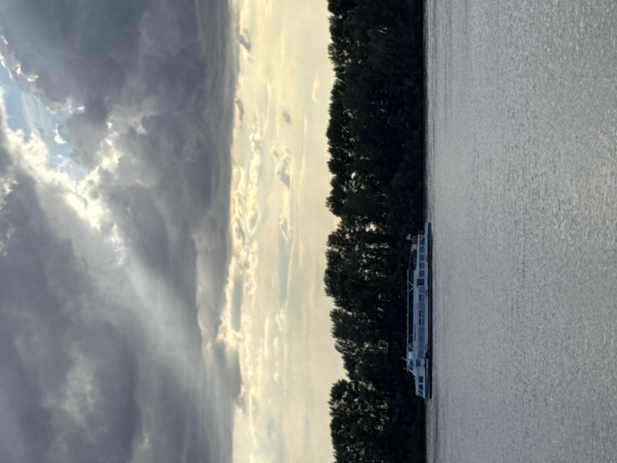 This is a vertical, low-angle, outdoor shot of a river with a boat and trees. The sky is filled with dramatic clouds, some dark and stormy, others bright and illuminated by the sun, suggesting it's either dawn or dusk. The sun's rays are breaking through the clouds, creating a dynamic and somewhat ominous atmosphere. On the right side of the frame, a dense line of dark green trees runs along the riverbank, their leaves creating a textured silhouette against the bright sky. In the water, close to the trees, a long, white passenger boat is visible. It appears to be moving slowly or is docked, with multiple decks and windows. The water itself is a muted silver-grey, reflecting the sky and giving a sense of calm despite the turbulent clouds. The overall impression is one of natural beauty and serene movement on the water, with the dramatic sky adding a touch of drama. There is no visible text in the image. The scene evokes a sense of peaceful journey or quiet observation in nature.