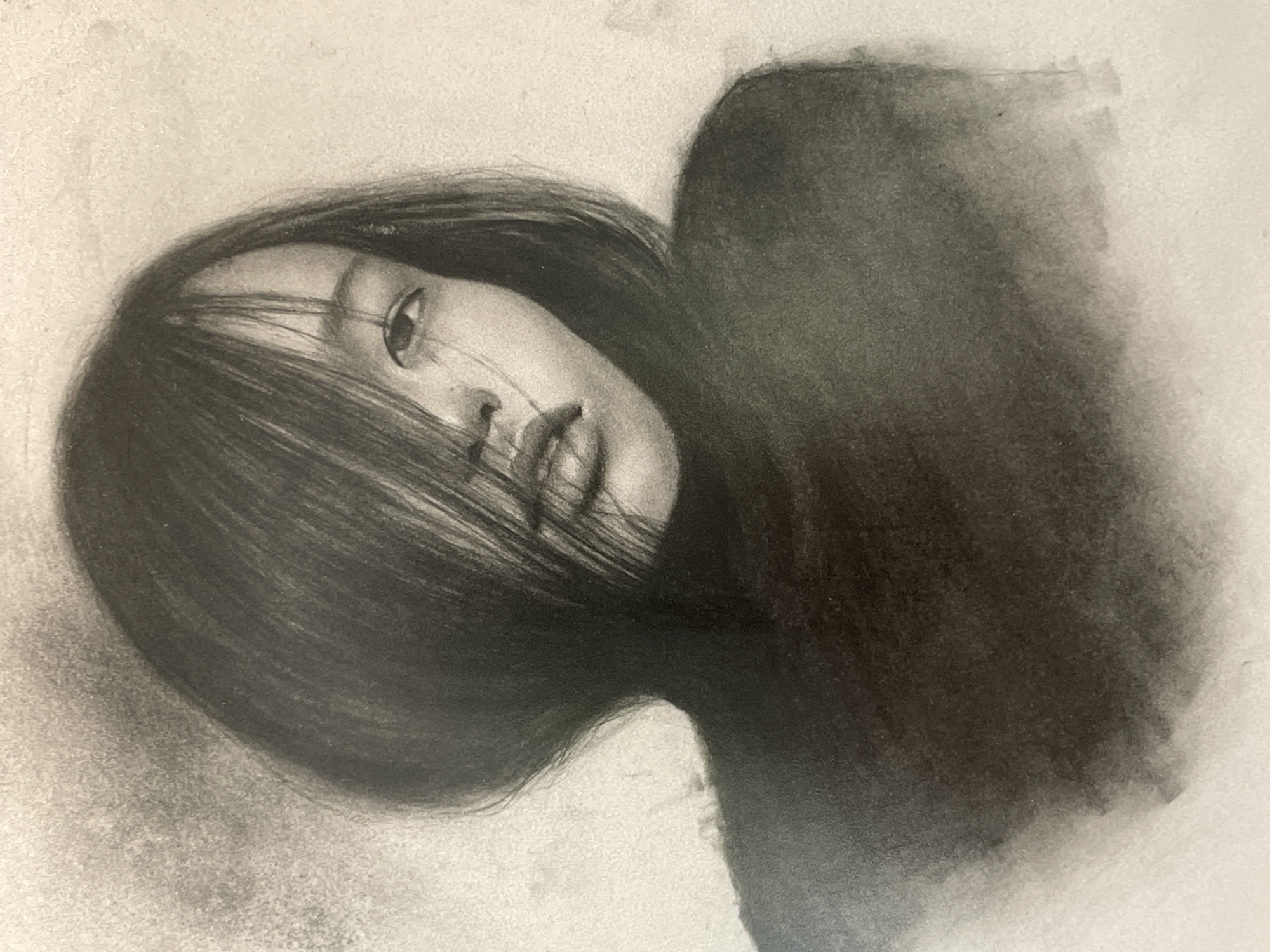 A charcoal drawing depicts a close-up portrait of a young Asian woman. Her head is tilted to the side, with her dark hair partially obscuring her face. One eye is clearly visible, with delicate shading around it. Her lips are slightly parted, conveying a subtle expression. She is wearing a dark, high-necked garment. The background is a textured, light grey wash, fading into white. The overall tone of the drawing is somber and evocative, with a strong emphasis on light and shadow.