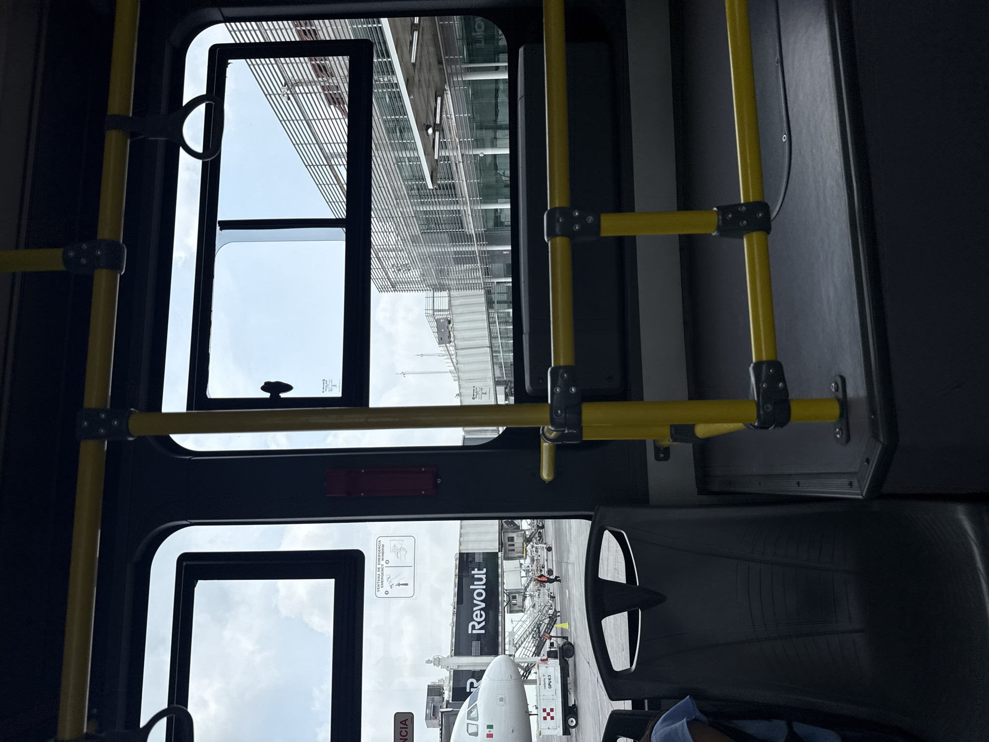 This image captures a scene from inside a public transport vehicle, likely an airport shuttle bus, looking out through its windows. The photograph is oriented vertically, showing elements both inside and outside the bus.
**Subjects & Scene:** Inside the bus, prominent yellow grab bars with black connectors stretch across the frame, indicating a typical public transport interior. Dark interior panels and a portion of a dark seat are visible in the lower right. Outside the upper window, a modern airport terminal building with a facade of glass and horizontal slatted structures can be seen under a cloudy sky. Outside the lower window, an active airport apron is visible. The front section of a white airplane, displaying what appears to be a Mexican flag, is a central feature. A jet bridge, clearly branded with the "Revolut" logo in white letters on a black background, is connected to the aircraft. Ground support equipment, including a cart with "AP-B303" printed on it, is positioned near the plane. An airport worker in an orange high-visibility vest stands on the tarmac.
**Activity:** The bus is stationary or moving slowly within an airport environment, transporting passengers or crew, or waiting on the apron. The airplane appears to be parked, possibly undergoing servicing or preparing for boarding/deplaning.
**Notable Details:** The scene takes place during the daytime, under a partly cloudy sky. Location cues such as the airplane, jet bridge, ground crew, and terminal building firmly place the setting at an airport, specifically in Apodaca, Mexico, as per the context.
**Visible Text:** "Revolut" is clearly visible on the jet bridge. On one of the bus windows, "VENTANA DE EMERGENCIA" (Emergency Window) and a corresponding instruction diagram are present. Text "AP-B303" can be seen on the ground support equipment.