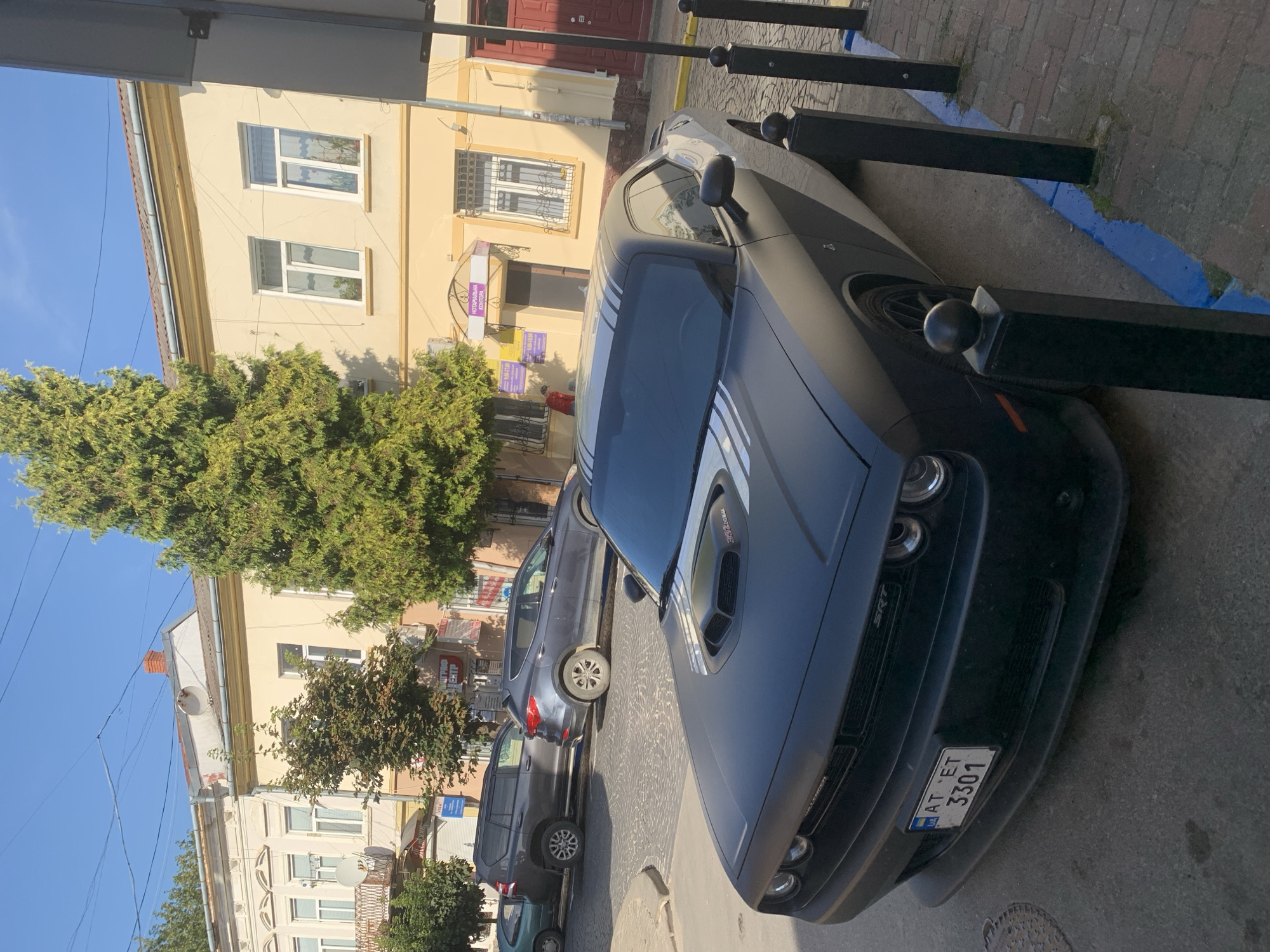 The image shows a black Dodge Challenger SRT parked on a cobblestone street in Kolomyia, Ukraine. The car is facing the viewer's left, with its front end prominently displayed. It has a matte black finish with silver racing stripes on the hood and roof. The license plate reads "AT ET 3301" with a Ukrainian flag symbol.
In the background, there are buildings with light yellow facades and tiled roofs. The architecture suggests an older European city. One building has the text "НОТАРІАЛЬНА КОНТОРА" above its entrance, indicating a notary office. Another building on the left has a balcony with an ornate metal railing.
Several other vehicles are visible in the background: a dark grey SUV and a smaller, older green car. Trees line the street, and utility wires are strung across the sky. The sun is shining, casting shadows and illuminating the scene.
The foreground is dominated by the car and several black bollards with spherical tops, demarcated by a blue line painted on the cobblestones. The overall impression is a street scene in a historic urban setting.