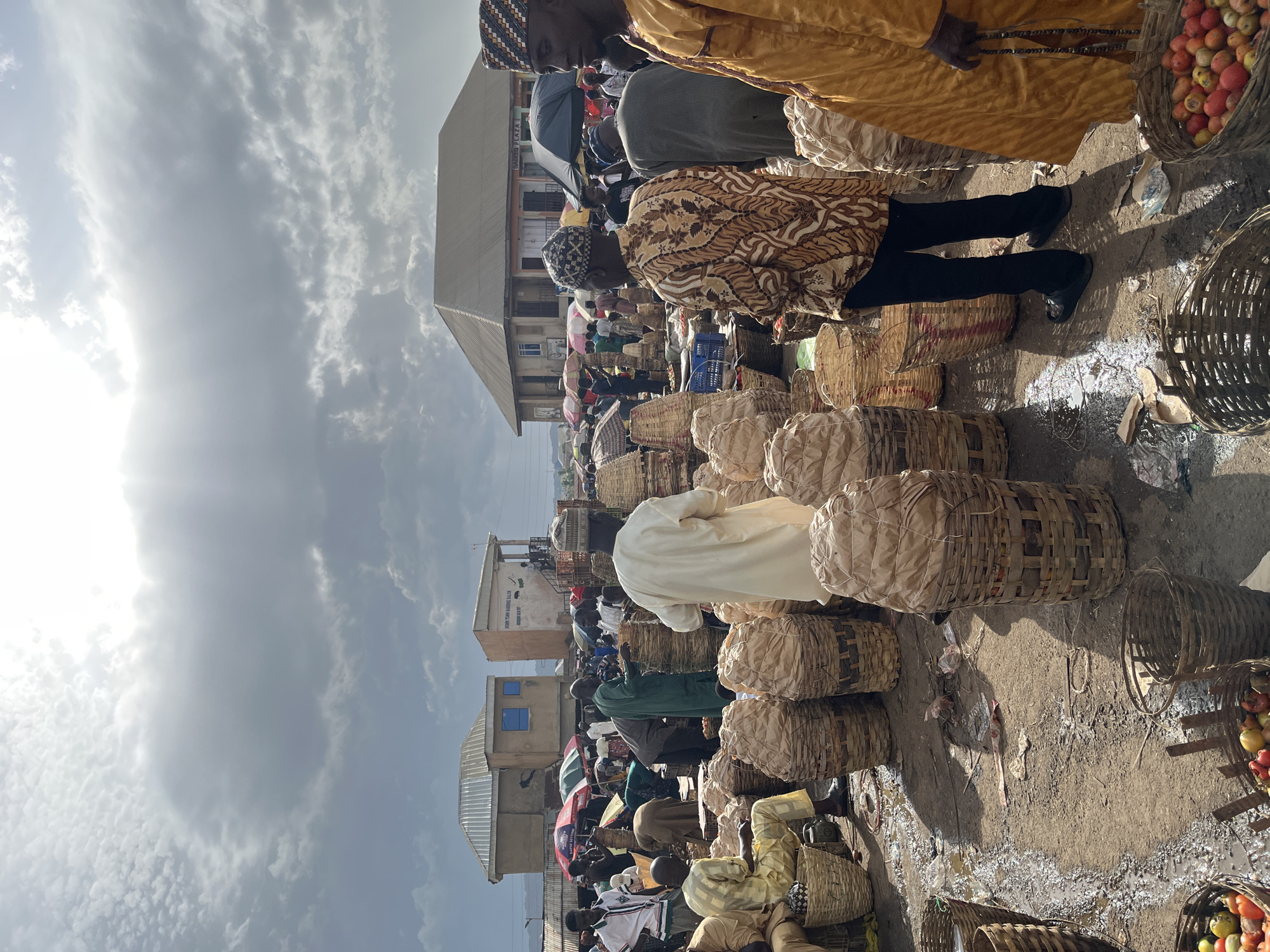 The image captures a bustling outdoor market scene, likely in Rafin Par, Nigeria, given the context provided by the crops. The sky is dramatic with a mix of sunlight breaking through dark, cloudy masses, suggesting changing weather conditions. In the foreground, large, woven baskets filled with what appears to be produce, possibly tomatoes and other fruits, are prominently displayed. Some baskets are covered with light-colored paper or cloth. Several men, dressed in traditional clothing including flowing robes and caps, are interacting within the market. One man in a white robe stands amidst the baskets, while another in a patterned brown and white shirt and black pants is also visible. A man in a bright yellow robe is seen from the side, holding prayer beads. The ground is dirt and appears wet in patches, with scattered debris. In the background, market stalls with umbrellas and various buildings, some appearing to be shops or residences with corrugated metal roofs, are visible, further indicating a lively commercial environment. The overall impression is one of vibrant market activity under a dynamic sky.