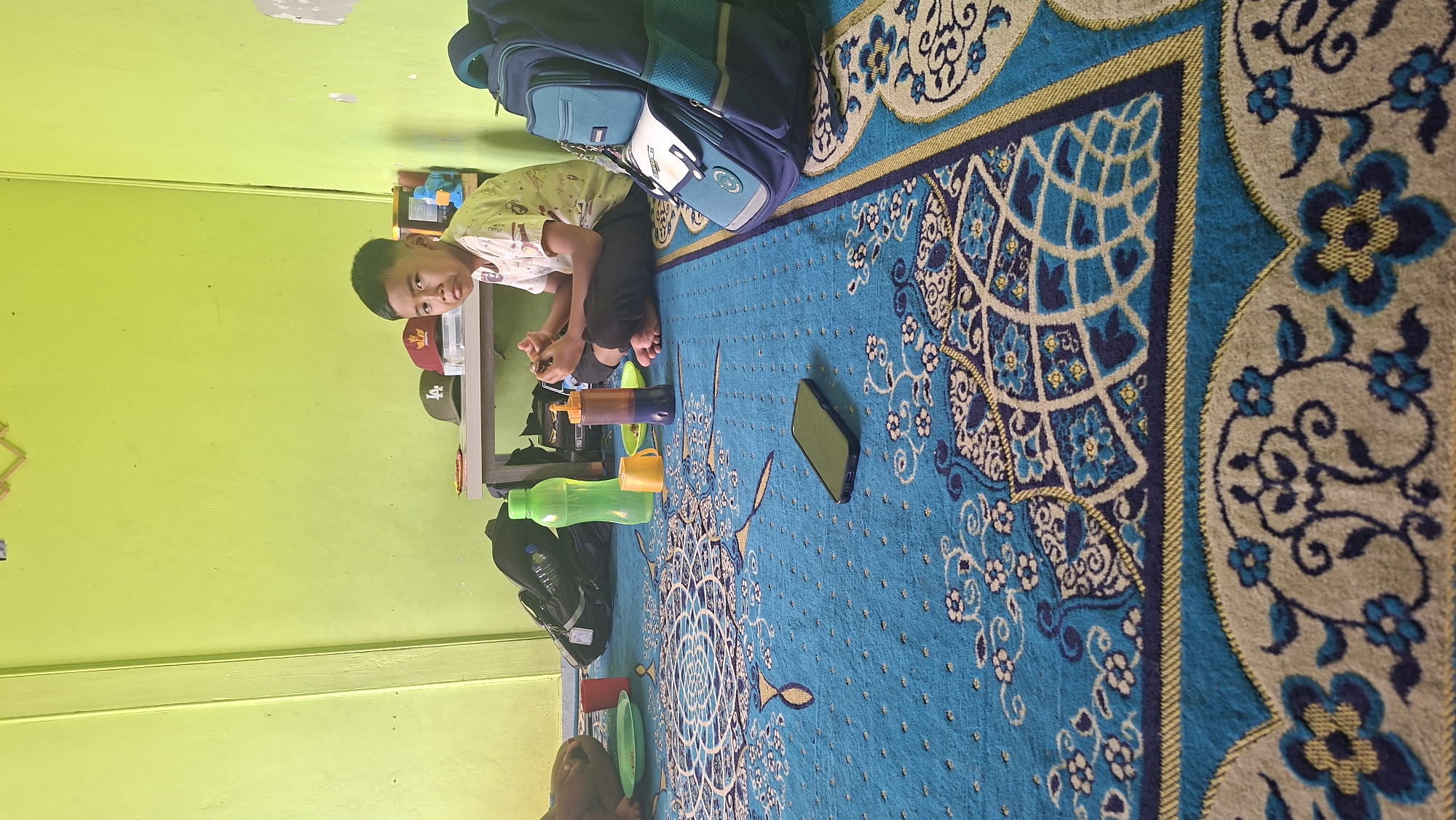 In Mlaten, Indonesia, a young boy sits cross-legged on a blue patterned carpet. He is wearing a light-colored t-shirt with a design and dark pants. In front of him, on the carpet, are a few objects: a smartphone, a red cup, and a yellow cup. To his right is a dark blue backpack with teal accents. In the background, a low table holds several items, including a green water bottle, another bottle with an orange dispenser, and several baseball caps. The wall behind the table is painted a bright lime green. The room appears to be a domestic interior.