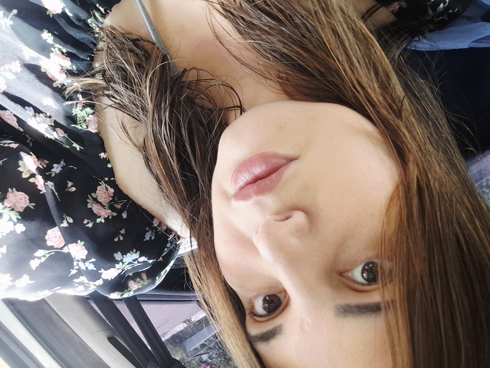 The image is a close-up, portrait-style selfie of a woman, possibly in her late teens or early twenties, with long, light brown hair that is wet and appears to be styled naturally. Her face is angled towards the camera, and her expression is neutral. She is wearing a dark floral-print top. The background suggests an indoor setting, possibly a car, with hints of blue fabric and what appears to be a seatbelt. The lighting is bright, with a slight shadow on her face. There is no indication of the city of Chaikovsky, Russia, or any specific urban environment.